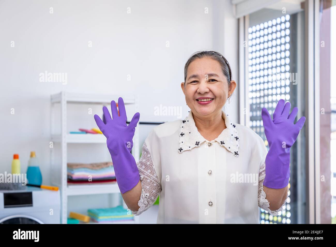 Donna anziana asiatica che indossa guanti di gomma e solleva le mani nella lavanderia con orgoglioso, guardando la macchina fotografica, concetto di pulizia primaverile Foto Stock