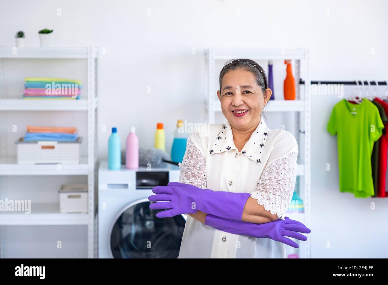 Donna anziana asiatica che indossa guanti di gomma e braccia incrociate nella lavanderia con orgoglioso, guardando la macchina fotografica, concetto di pulizia primaverile Foto Stock