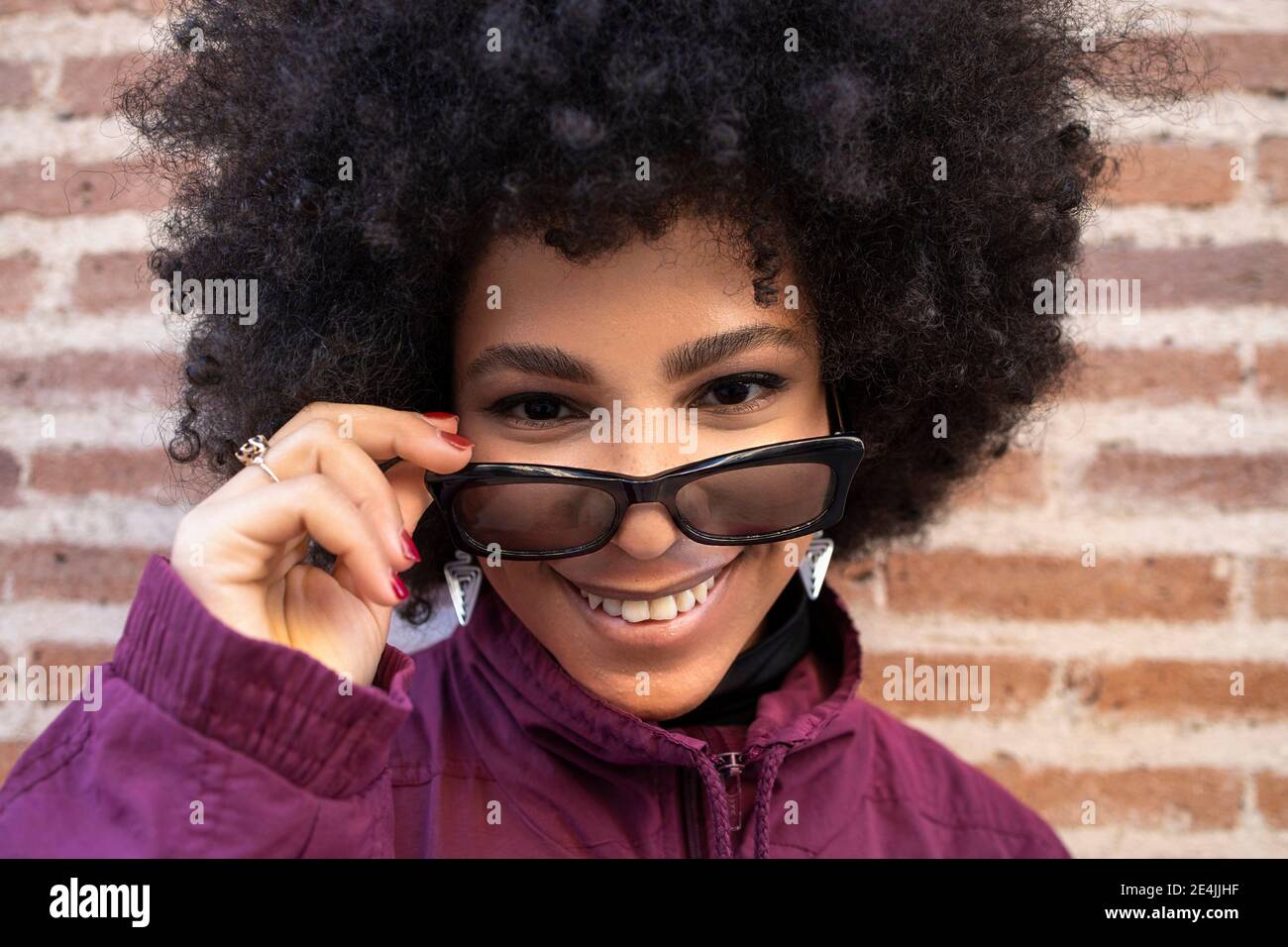 Afro donna con espressione vuota indossando occhiali da sole mentre in piedi contro muro di mattoni Foto Stock