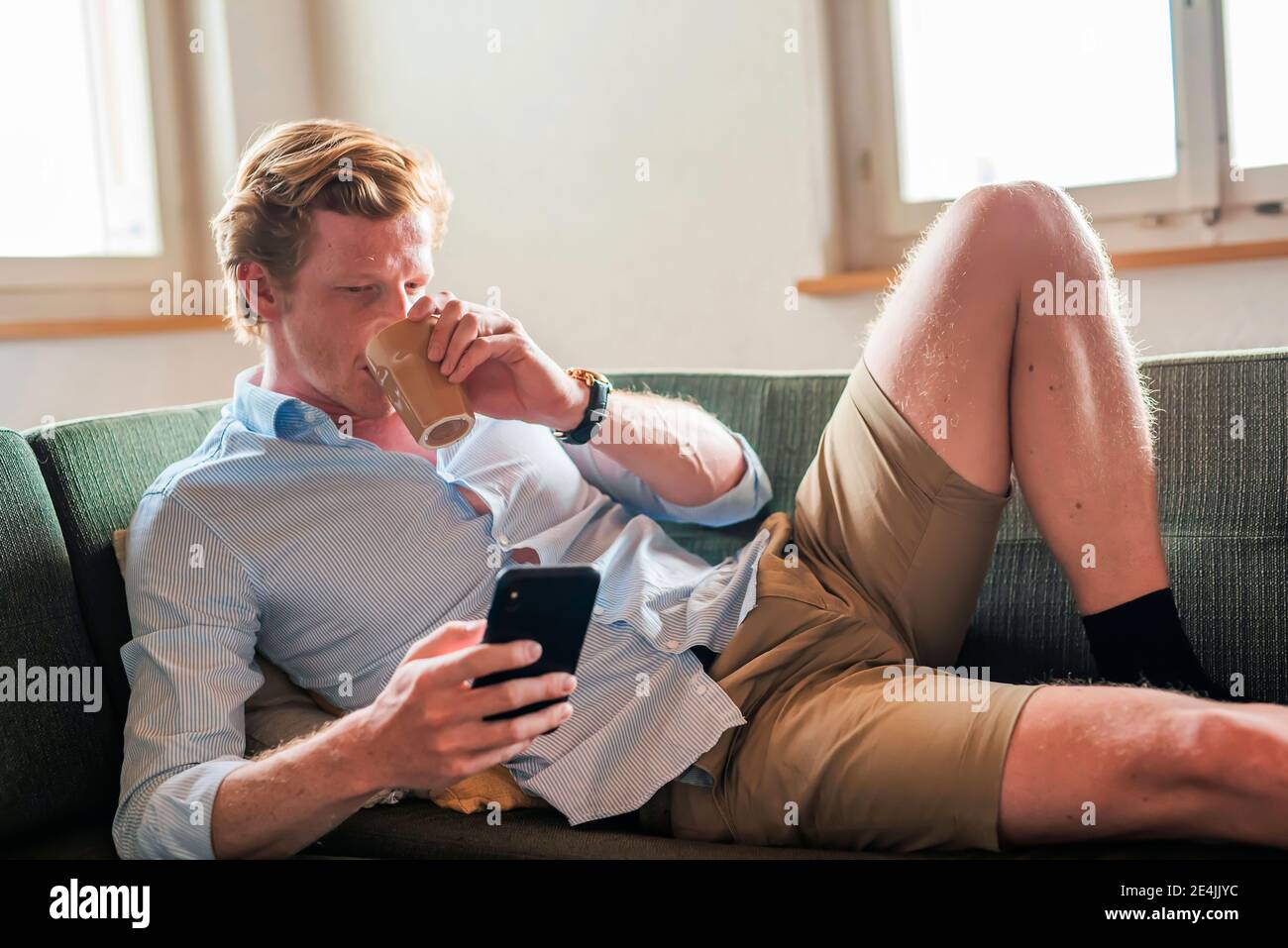 Uomo che beve caffè mentre usa lo smartphone in soggiorno Foto Stock