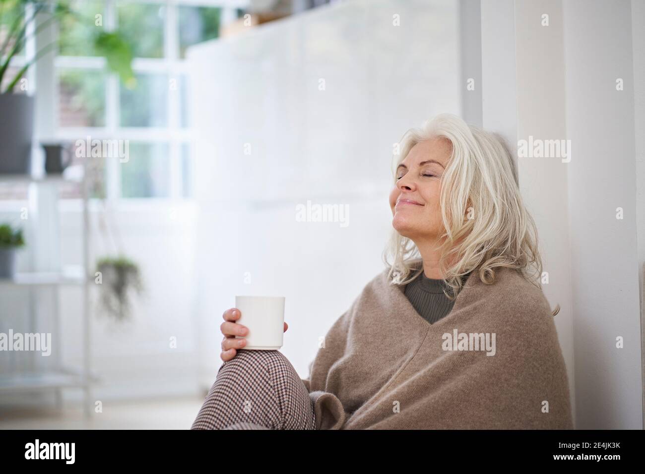 Donna anziana in pensione che tiene la tazza di caffè mentre si siede con gli occhi chiuso giorno sognando a casa Foto Stock