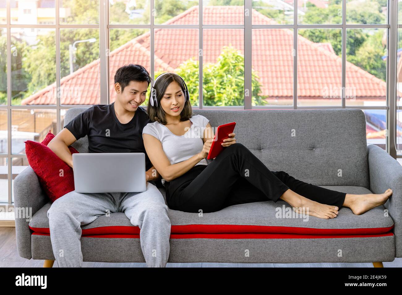 Giovane coppia asiatica rilassante seduta sul divano usando un computer portatile e tablet digitale che lavorano da casa Foto Stock