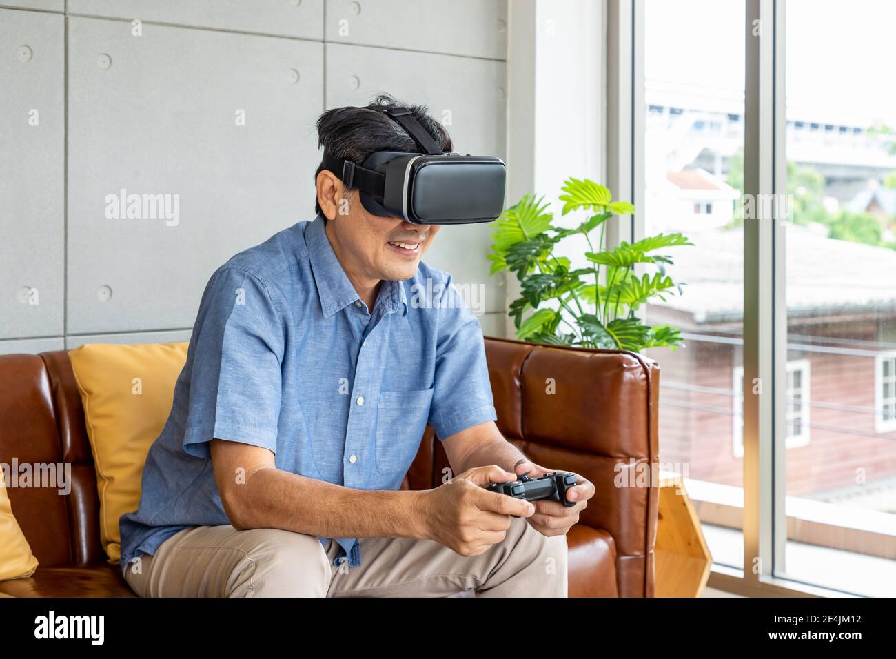 Anziano uomo asiatico di pensione che indossa i vetri di realtà virtuale seduto sopra divano che tiene il joystick e gioca Foto Stock