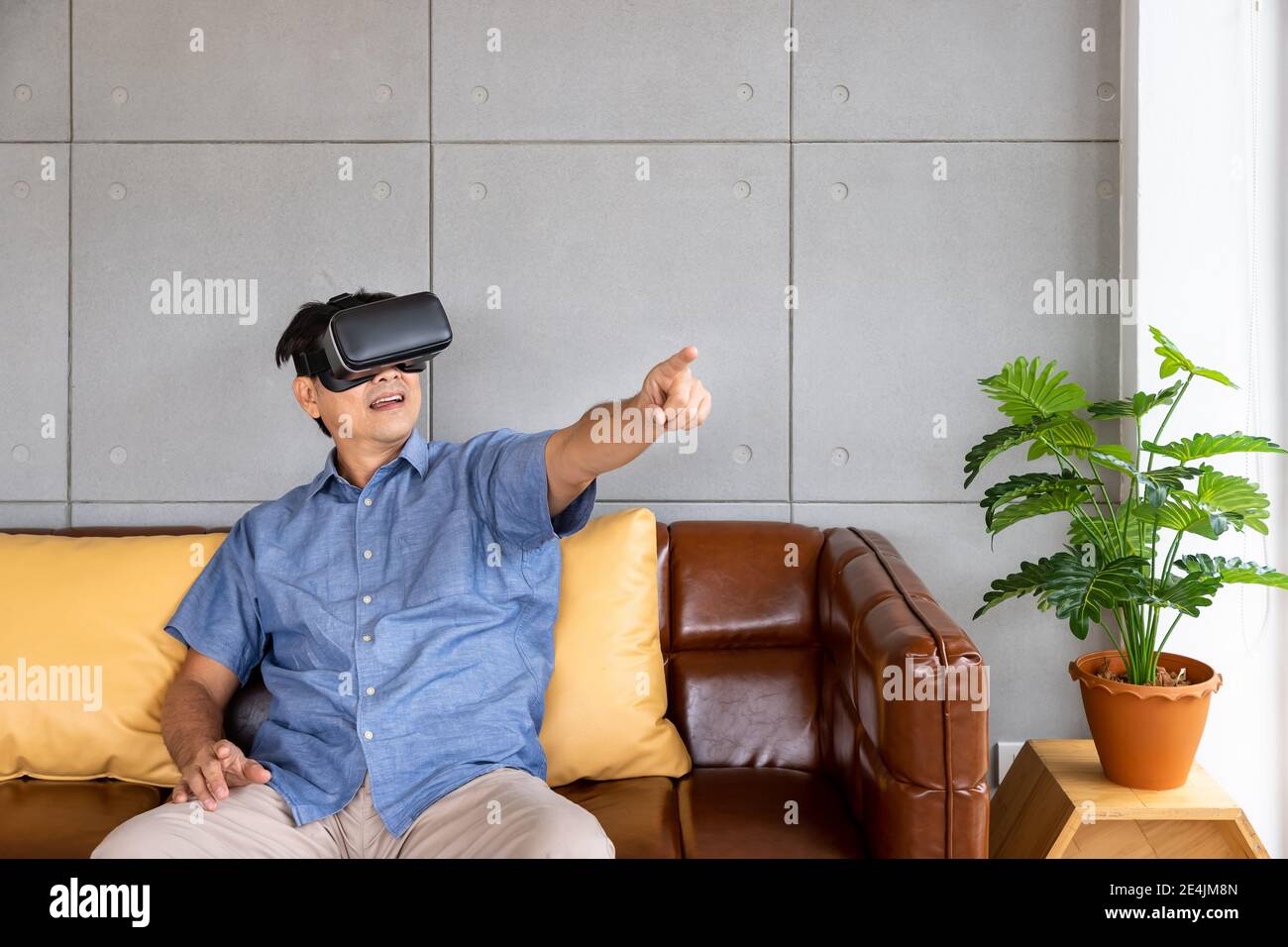 Anziano uomo asiatico di pensione che indossa i vetri di realtà virtuale seduto sopra divano goditi un'esperienza emozionante e puntando il pulsante Foto Stock