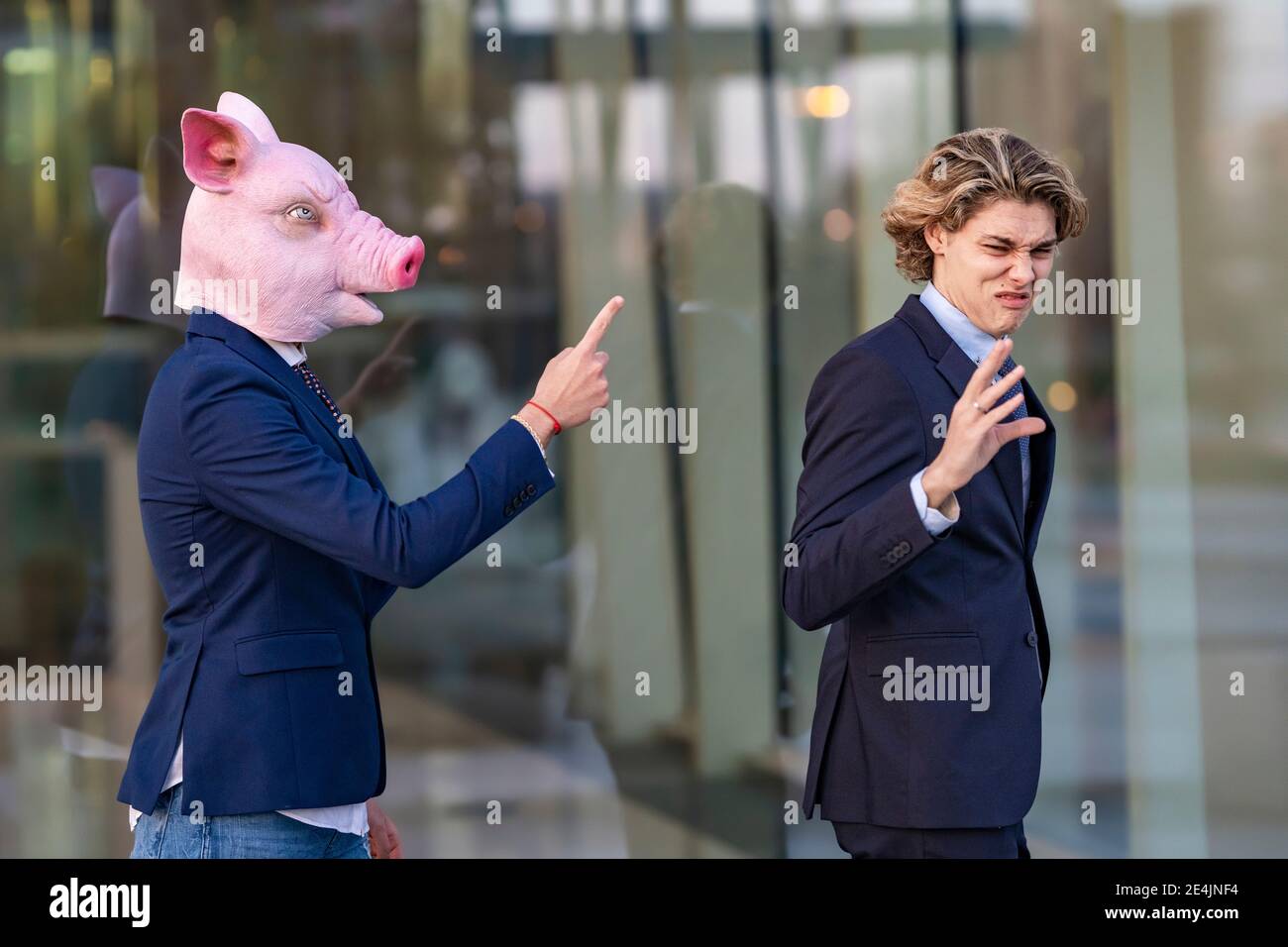 Giovane professionista maschile irritato da discutere con il collega in maiale maschera mentre camminando da parete di vetro Foto Stock