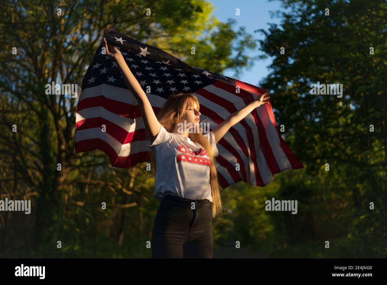 Donna bionda che tiene la bandiera americana mentre in piedi con gli occhi chiusi alla foresta Foto Stock
