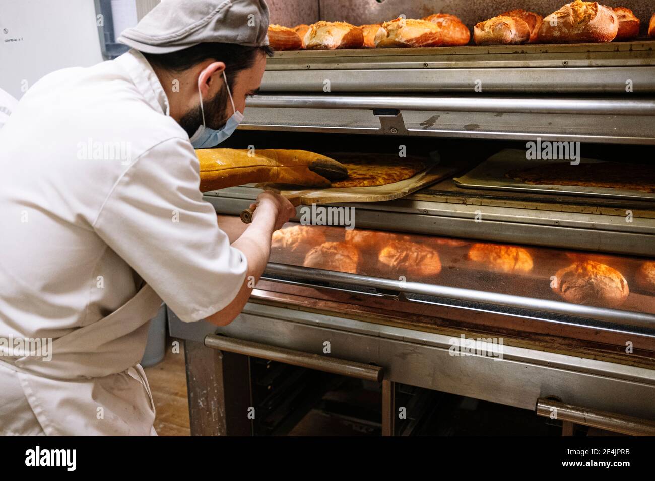 Panettiere maschio con buccia di pizza che controlla il pane in forno a. Panificio durante COVID-19 Foto Stock