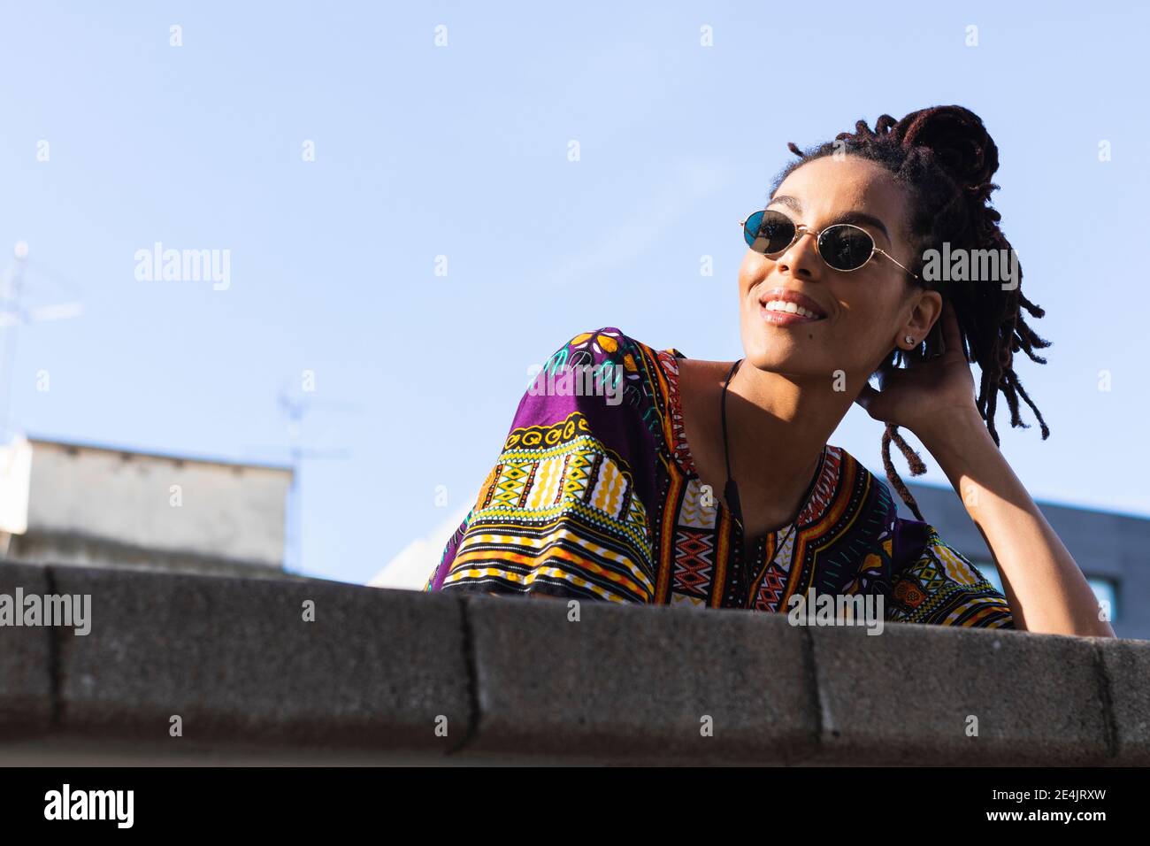 Donna con dreadlock che indossa occhiali da sole che guardano lontano mentre si appoggia parete di ritegno contro il cielo Foto Stock
