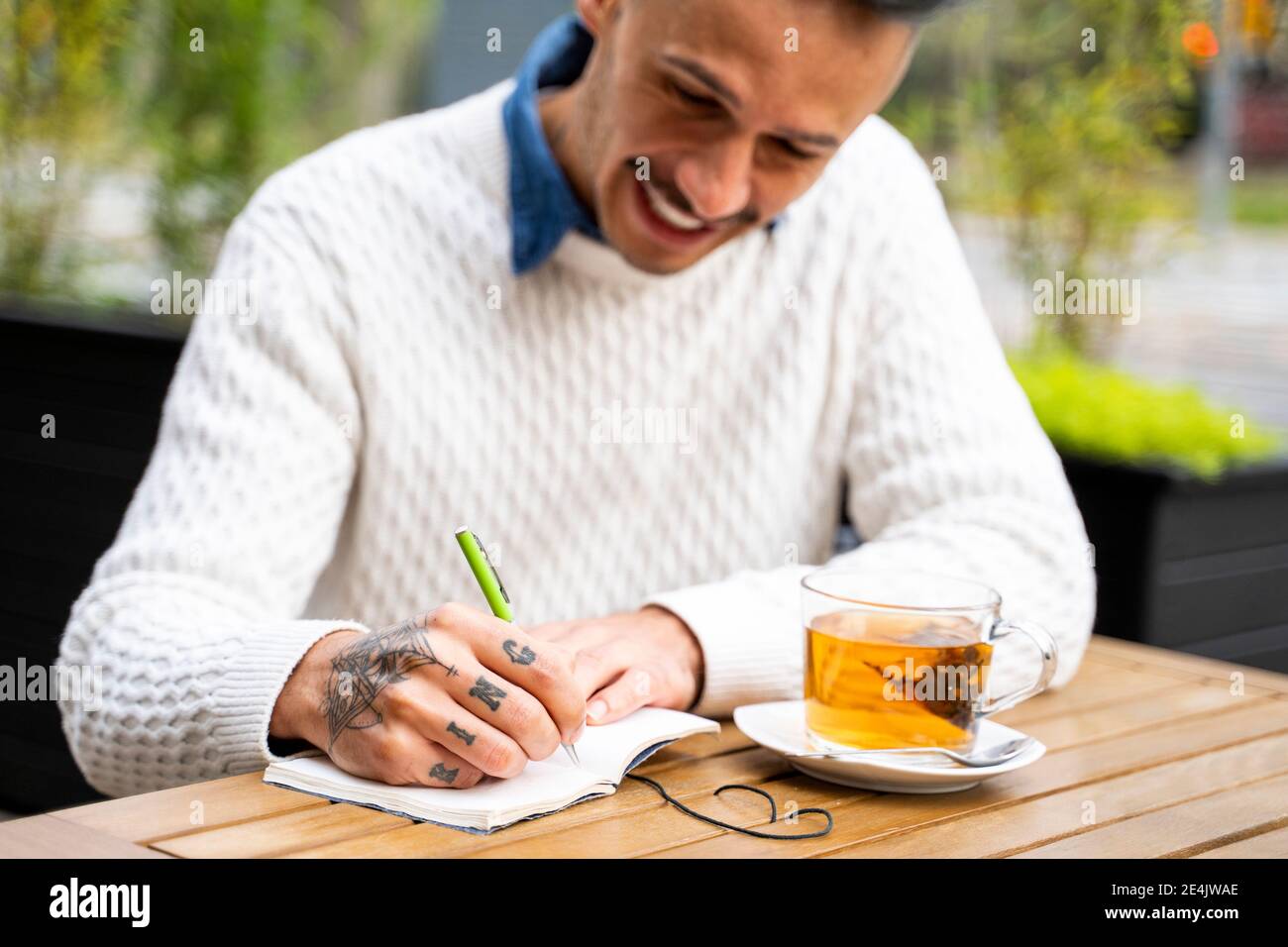 Uomo sorridente che scrive nel libro mentre prendendo la pausa del tè dentro bar sul marciapiede Foto Stock