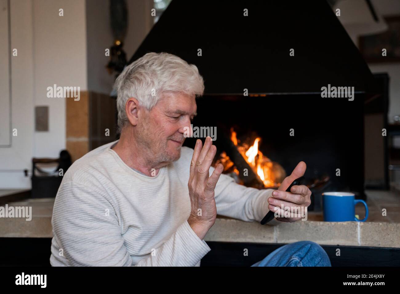 Uomo anziano che sventolava la mano per videochiamare sul telefono cellulare mentre si siede a casa Foto Stock