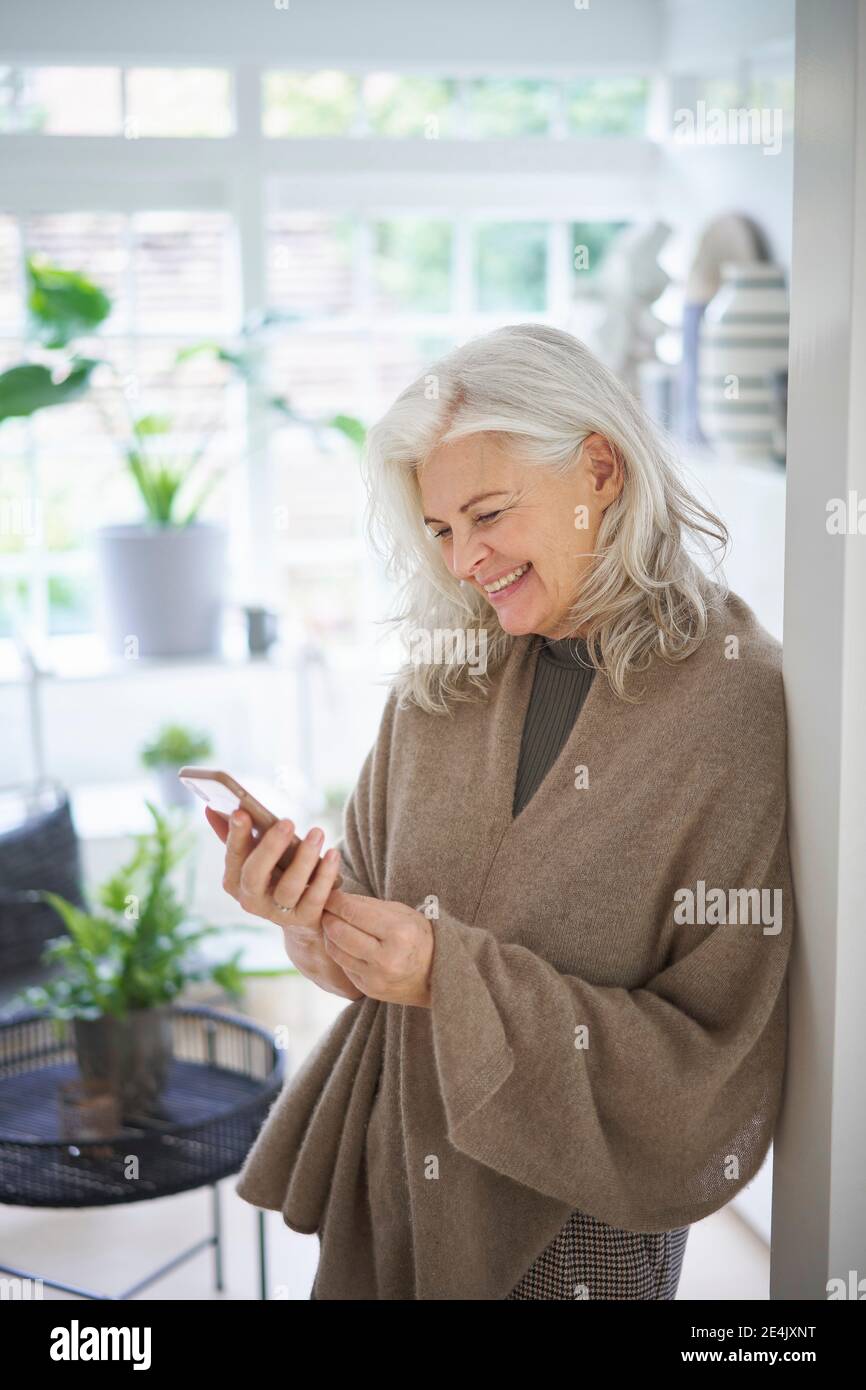 Sorridente messaggio di testo donna ritirata tramite smartphone a casa Foto Stock