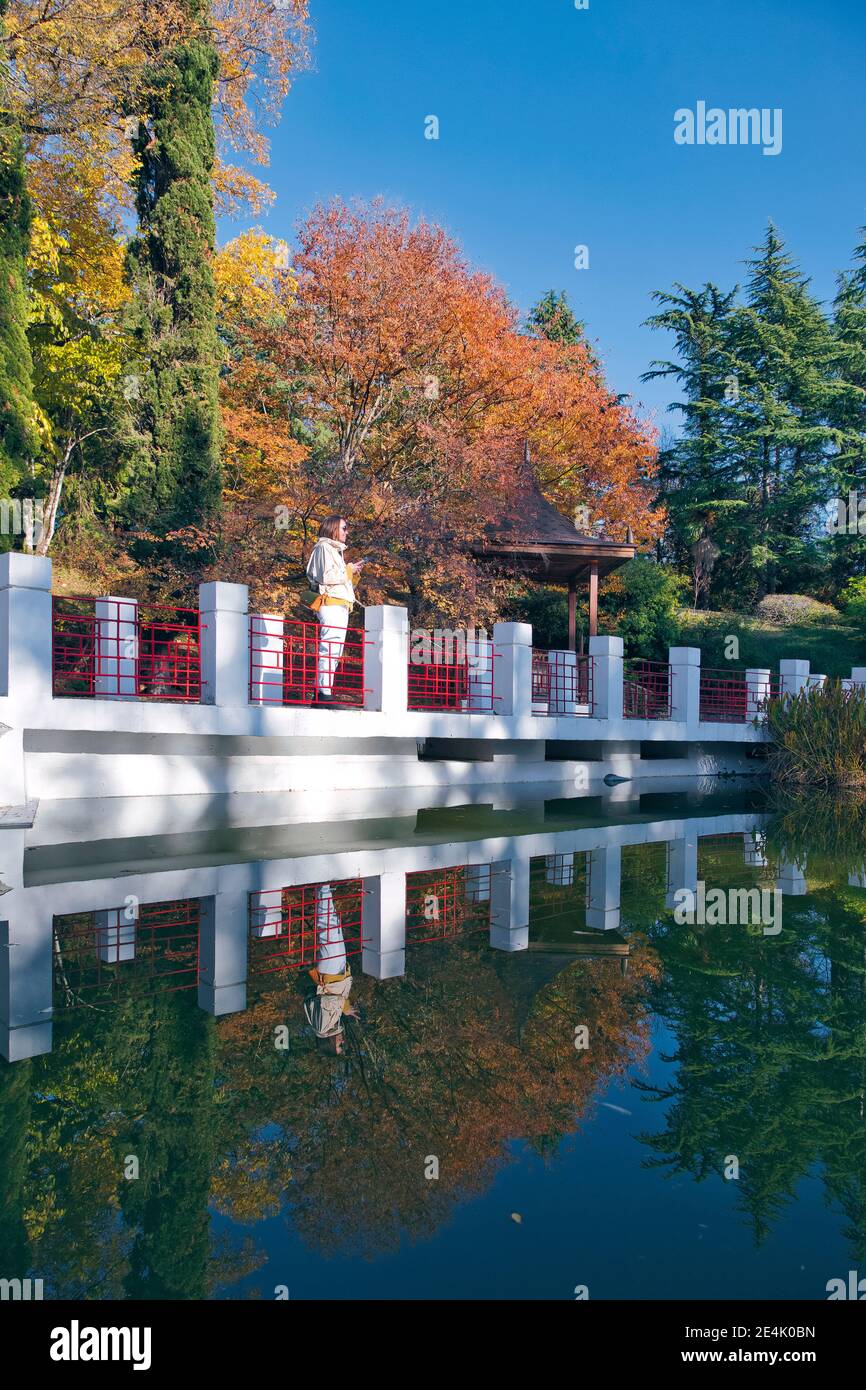 Russia, Krasnodar Krai, Sochi, giovane donna in piedi dietro recinzione al bordo dello stagno nel parco autunnale Foto Stock