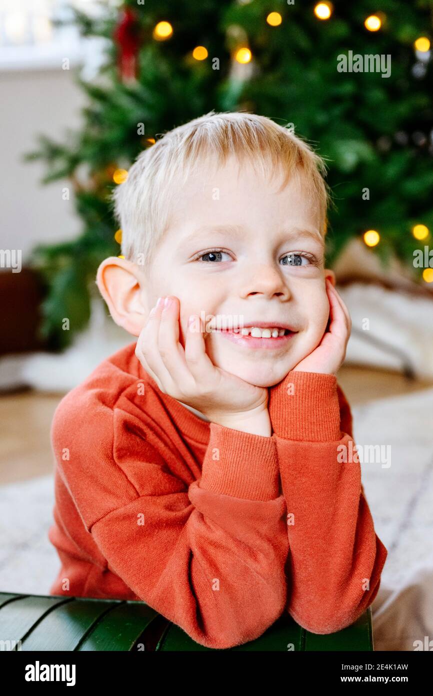 Primo piano ritratto del ragazzo carino con le mani sul mento seduto Con albero di Natale in background Foto Stock