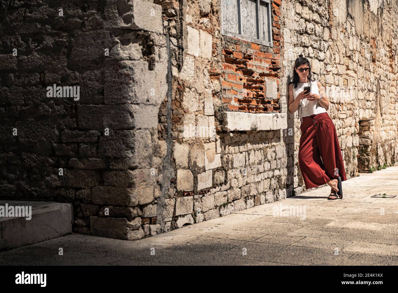Donna appoggiata contro il vecchio muro di pietra e guardando smartphone Foto Stock