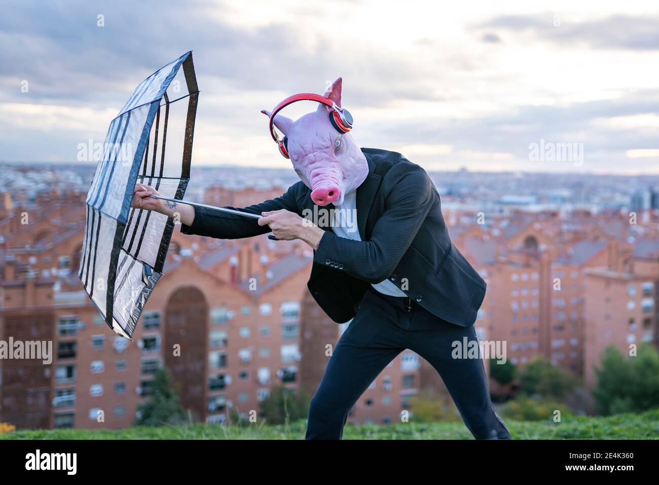 Imprenditore maschile indossando maschera di maiale e cuffie wireless che lottano con ombrello contro il cielo Foto Stock