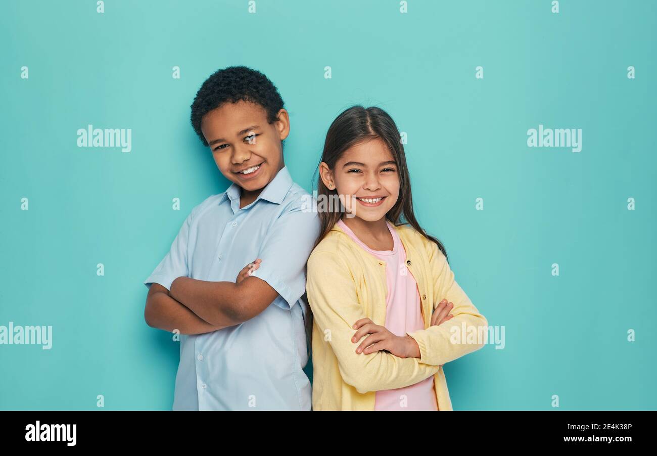 Ragazza asiatica e ragazzo afroamericano in piedi indietro con braccia incrociate su sfondo turchese Foto Stock