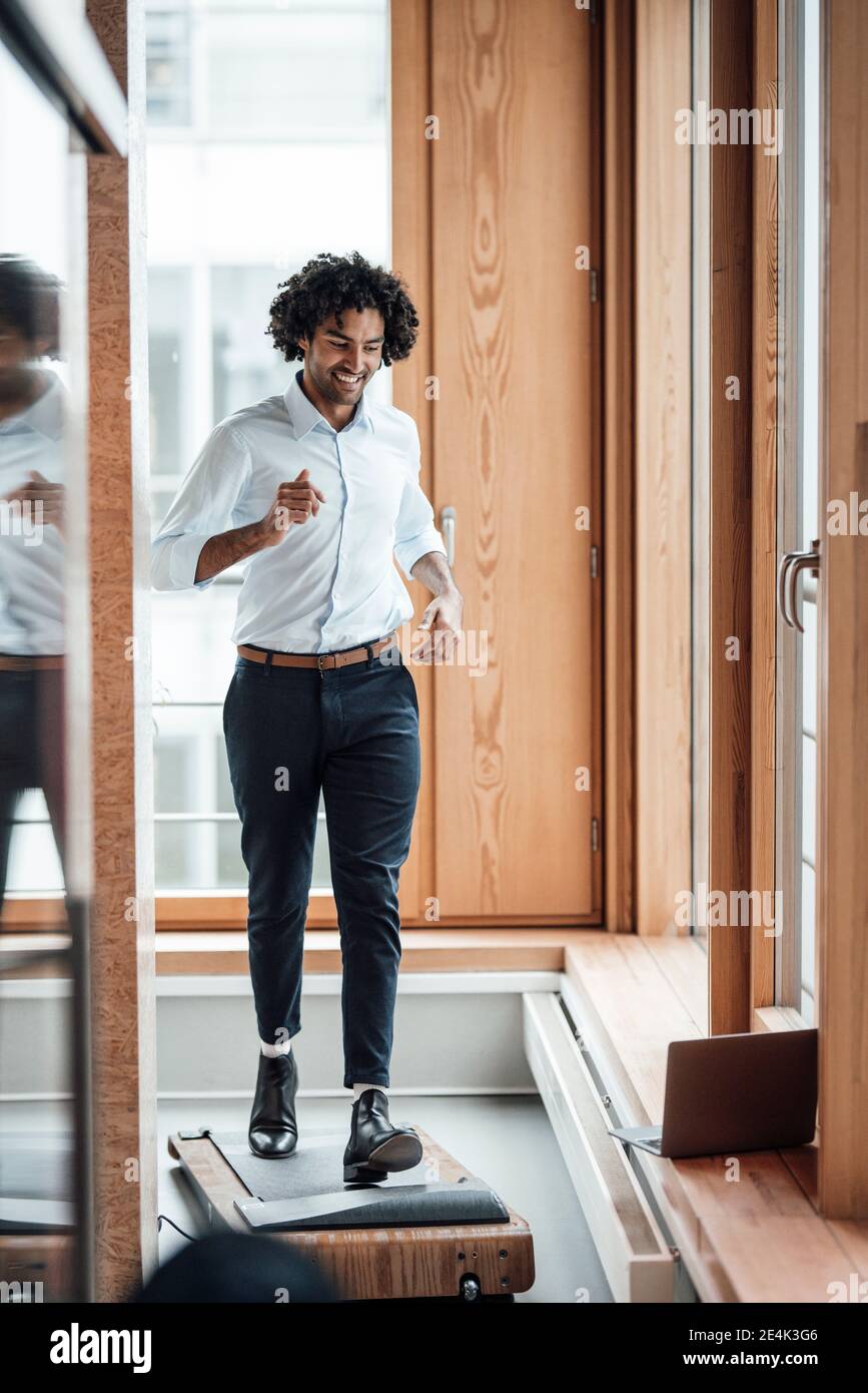 Sorridente imprenditore maschile attivo che fa jogging sul tapis roulant contro la finestra in luogo di lavoro Foto Stock