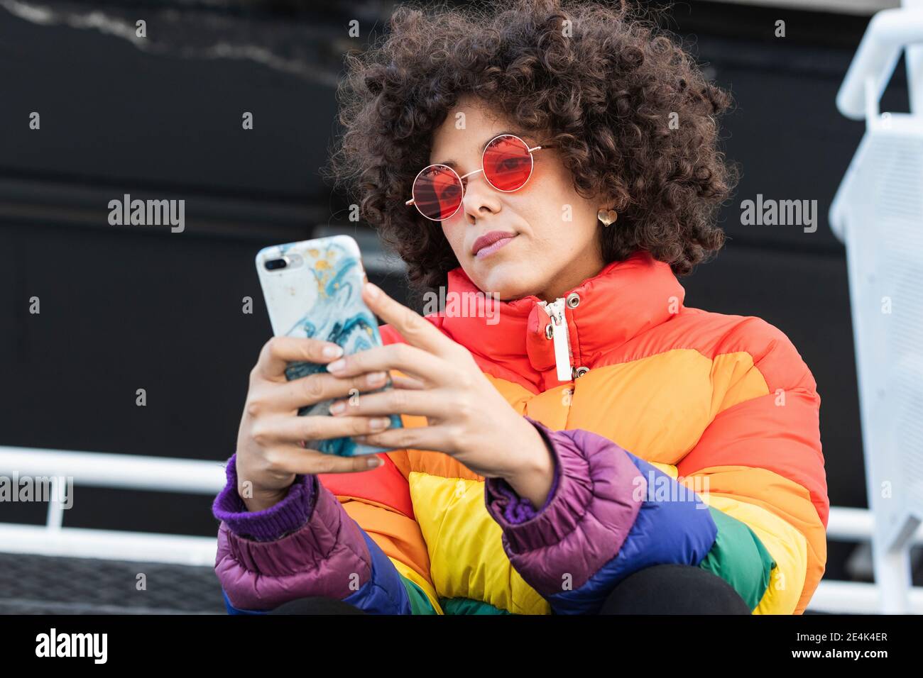 Donna con capelli ricci che indossa occhiali da sole e giacca calda usando il cellulare telefono mentre si è seduti all'aperto Foto Stock