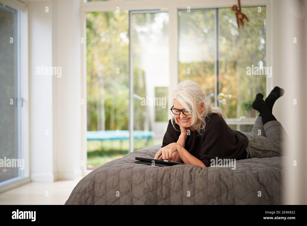 Donna anziana sorridente che usa il tablet digitale mentre si sdraiava a letto a casa Foto Stock