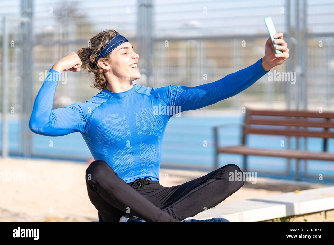 Sorridente atleta maschio che mostra bicep mentre prendendo selfie pubblico parcheggia il giorno di sole Foto Stock