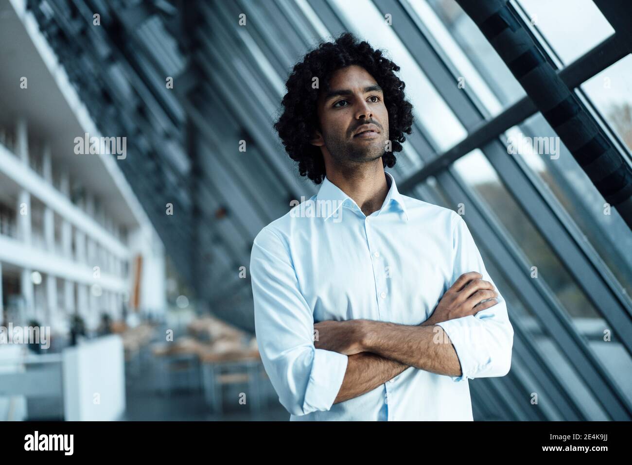 Pensieroso giovane uomo d'affari in piedi con le braccia incrociate mentre si guarda via sul posto di lavoro Foto Stock