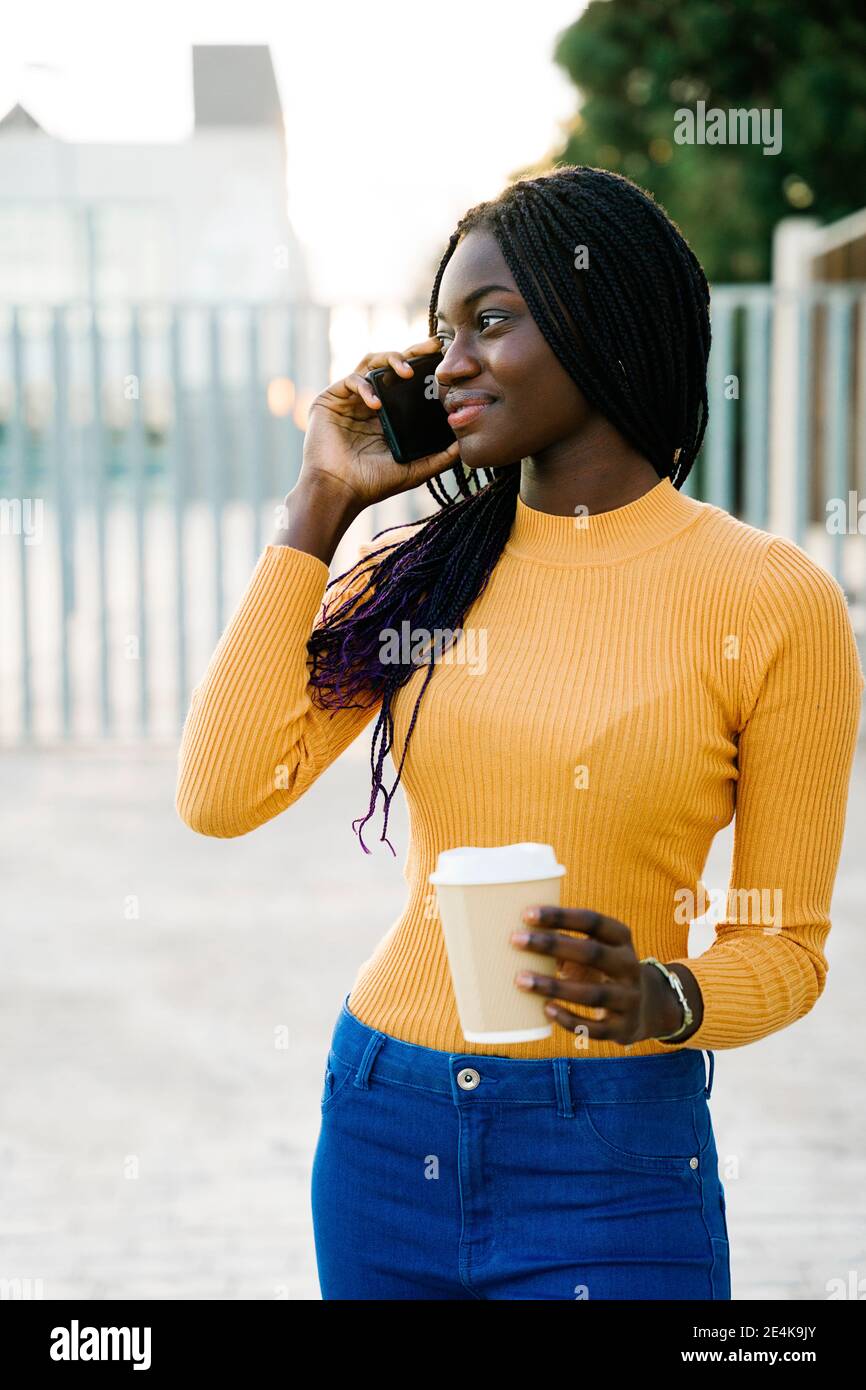 Sorridente ragazza adolescente con tazza di caffè guardando via mentre parla su smartphone in strada Foto Stock