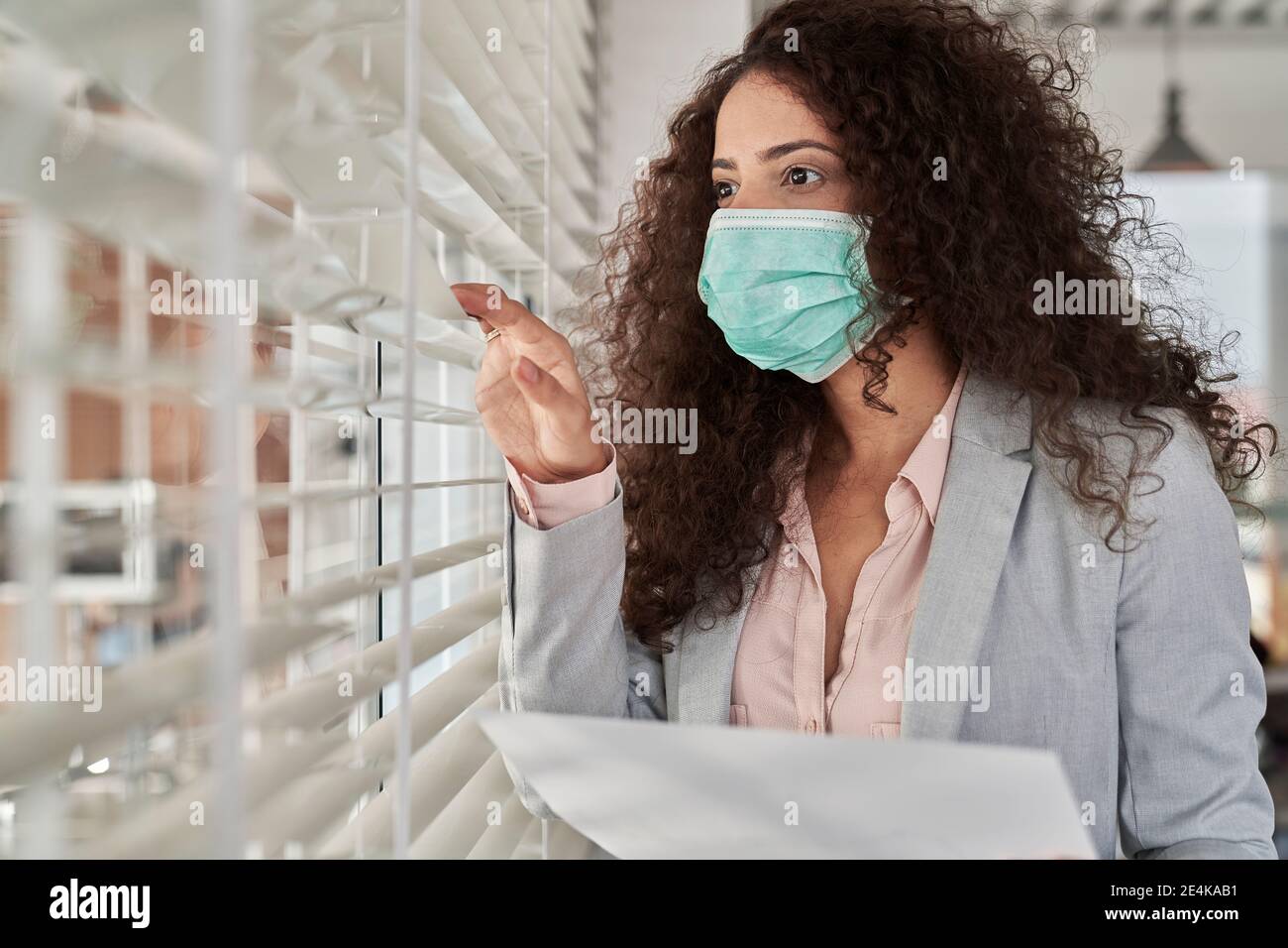 Giovane donna d'affari che indossa maschera protettiva che guarda attraverso le persiane sul posto di lavoro Foto Stock