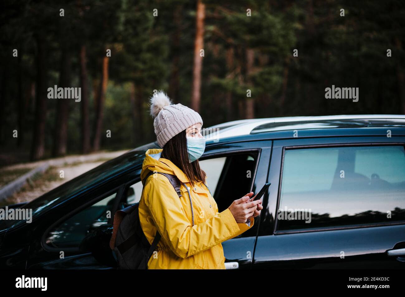 Donna che indossa un impermeabile giallo e una maschera facciale in piedi vicino all'auto, tenendo in mano uno smartphone Foto Stock