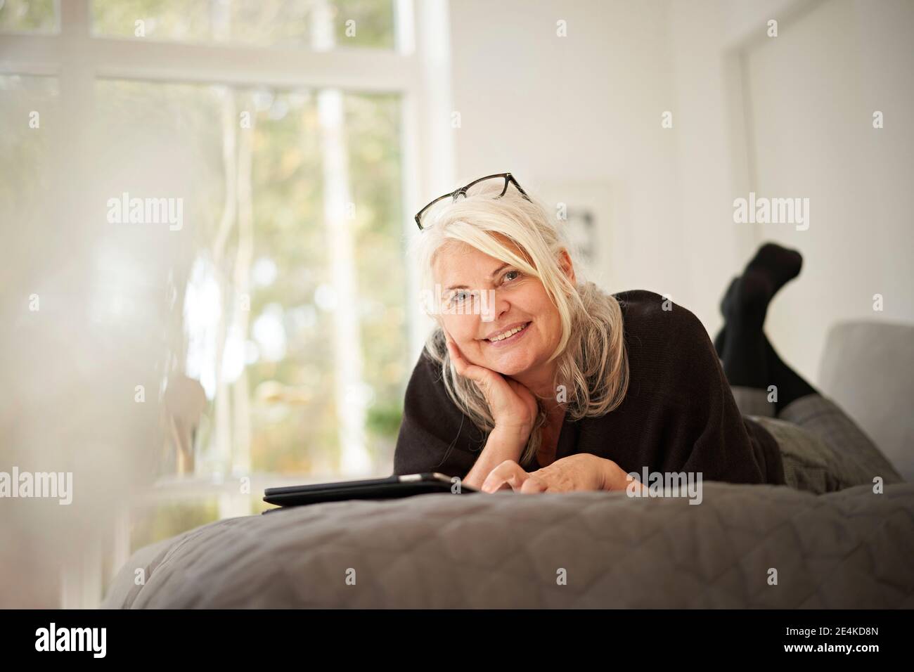 Donna anziana sorridente con la mano sul mento sdraiata sul letto a casa Foto Stock