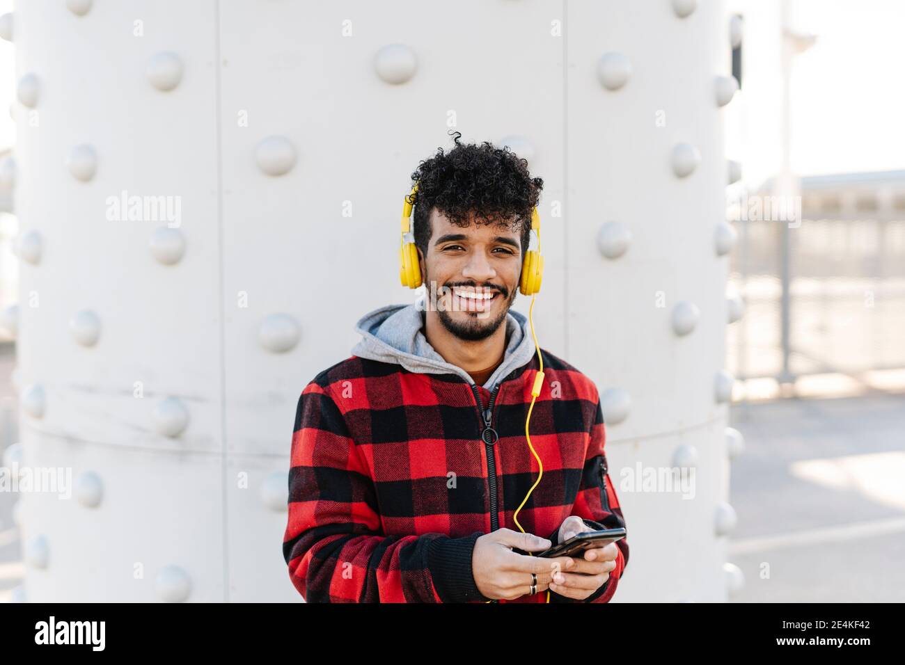 Uomo che indossa una camicia con cappuccio per ascoltare la musica attraverso le cuffie mentre si è in piedi con il telefono cellulare contro la parete Foto Stock