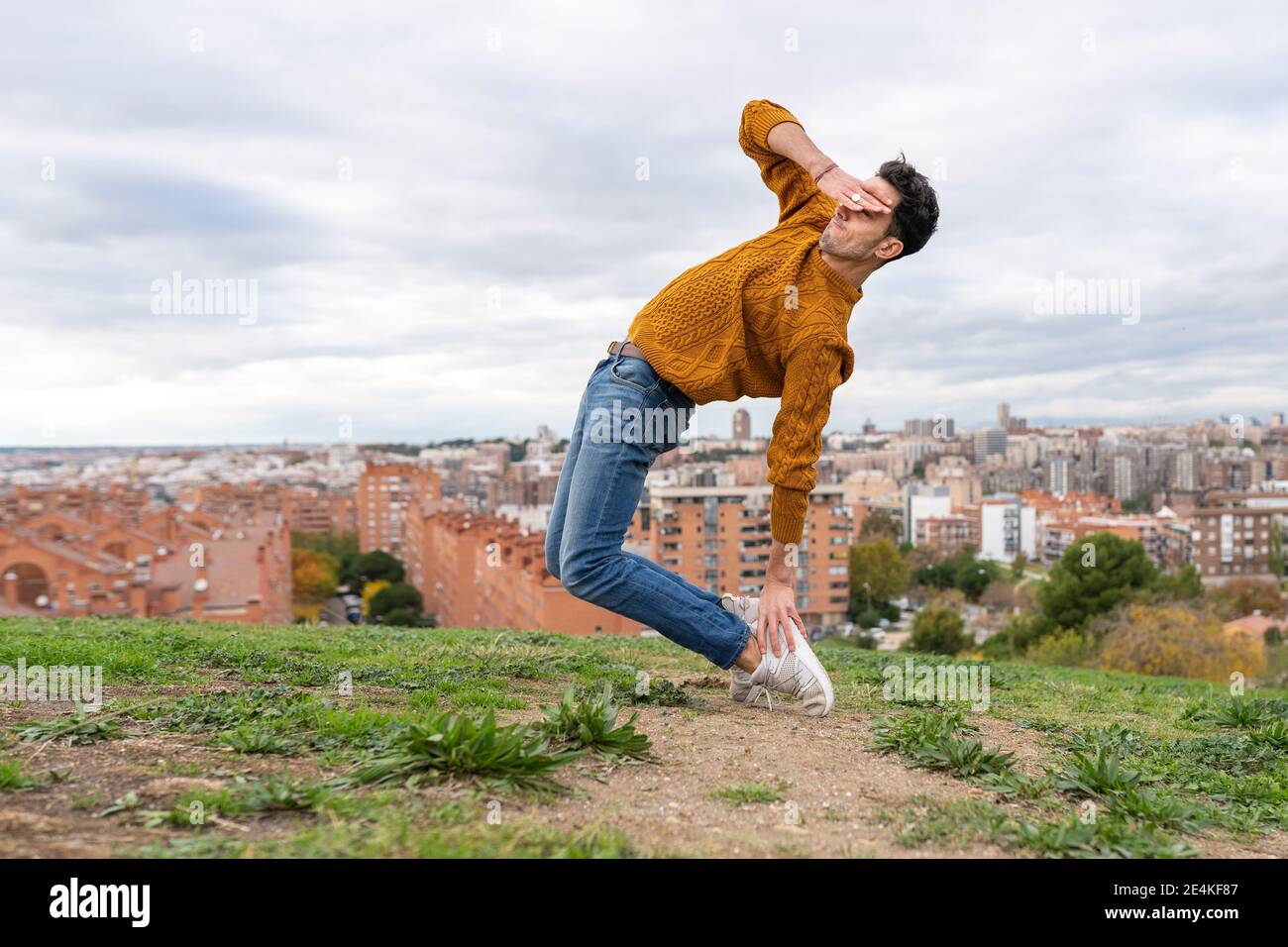 Flessibile giovane uomo scherma gli occhi mentre fa attività acrobatica su collina in città contro cielo nuvoloso Foto Stock