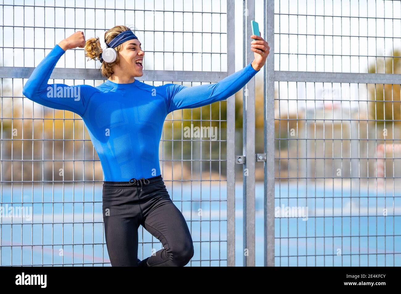 Atleta maschile con bocca aperta prendendo selfie mostrando bicep contro recinzione in giornata di sole Foto Stock