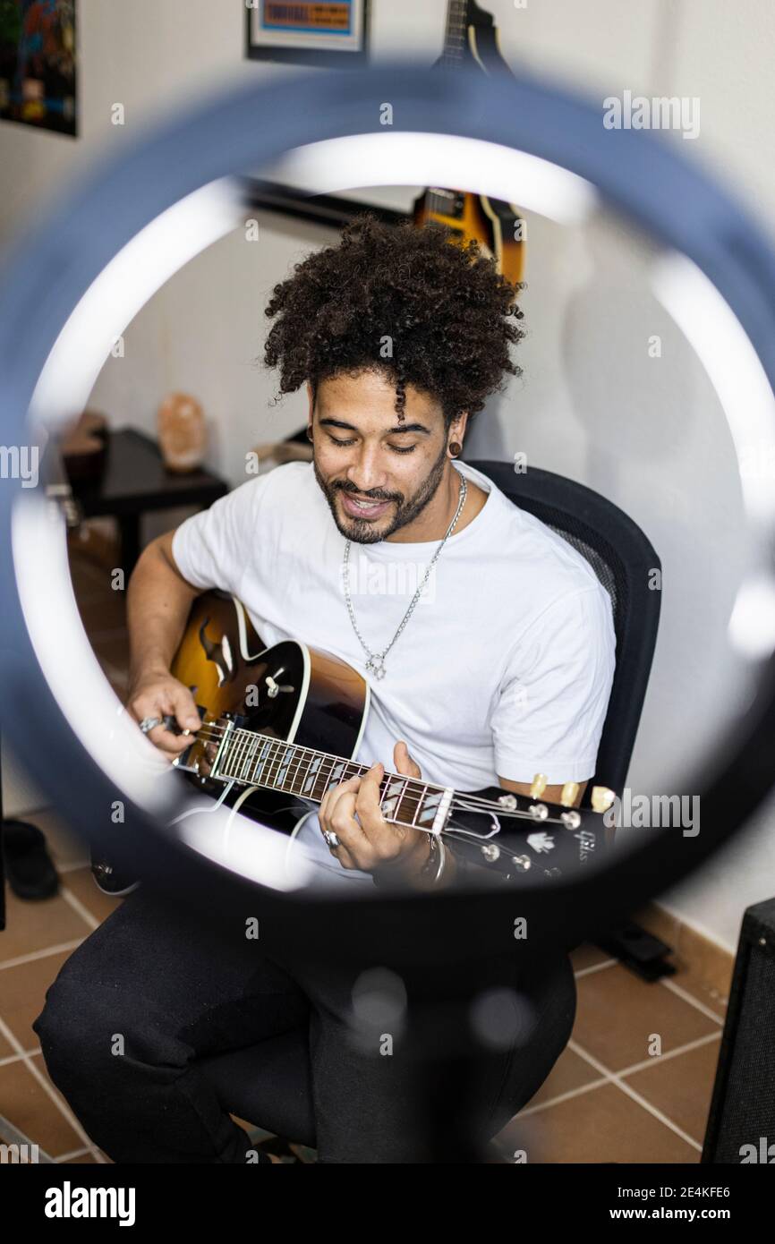 Sorridente giovane con webcam leggera che suona la chitarra mentre si è seduti a casa Foto Stock
