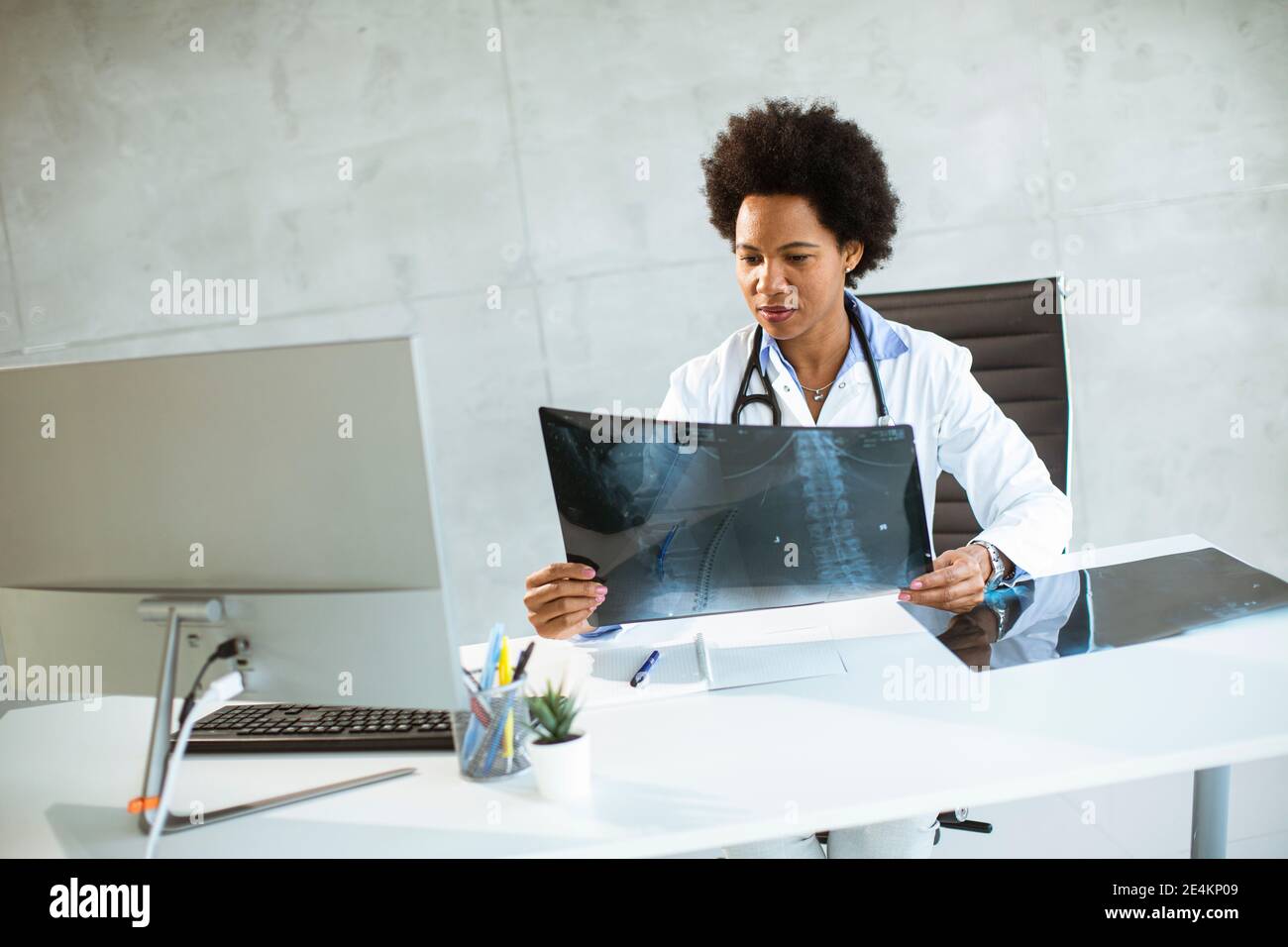 Donna Africana americana medico indossando camice bianco con stetoscopio seduta dietro la scrivania in ufficio e guardando l'immagine radiografica Foto Stock