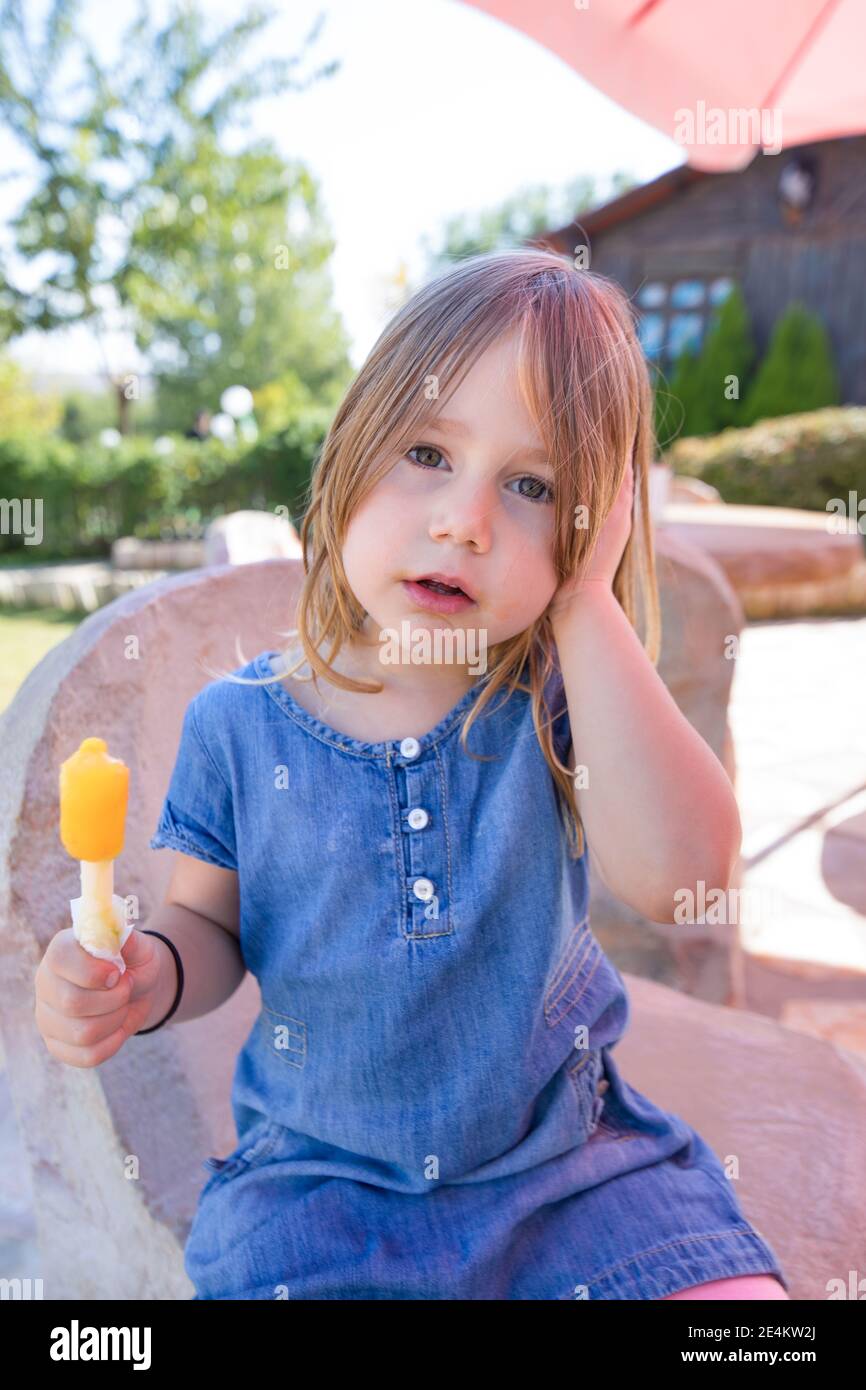 ritratto di una bambina bionda di quattro anni che guarda con un abito in denim blu, tenendo in mano lecca di ghiaccio giallo o arancione colorato, seduto in un parco pubblico Foto Stock
