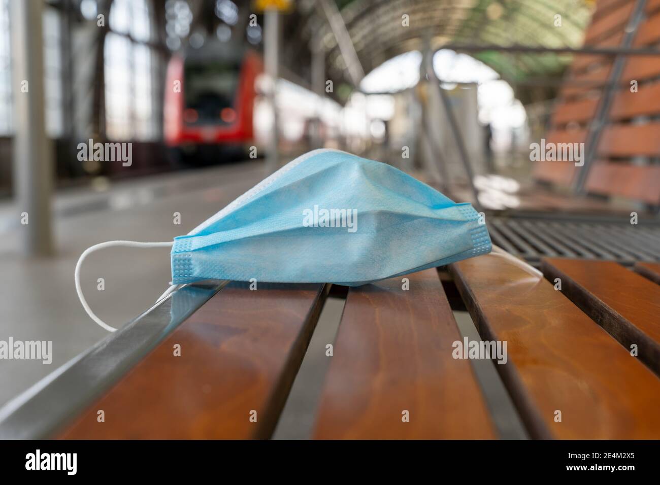 Maschera medica su una panchina in un'area di attesa di una piattaforma ferroviaria in una stazione. Protezione Covid monouso con un treno rosso sullo sfondo. Foto Stock