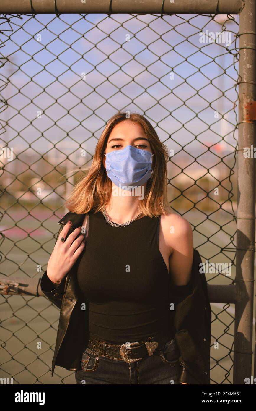 Ritratto di una ragazza adolescente con una maschera medica viso sulla strada, guardando la macchina fotografica e appoggiandosi ad una recinzione. Foto Stock