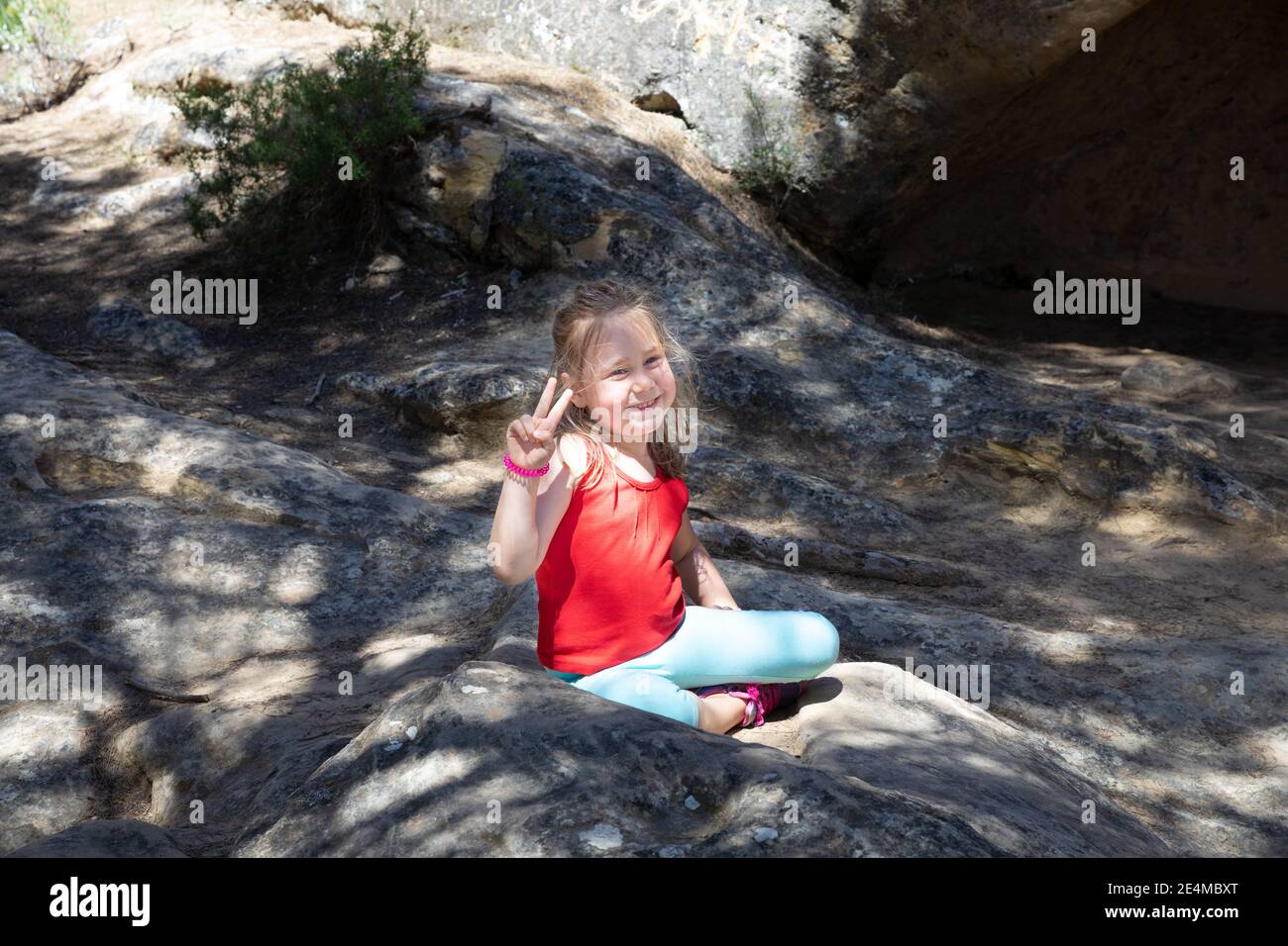 ritratto di quattro anni bionda carina ragazza, seduta sulla roccia all'aperto, guardando e facendo con le dita di mano il simbolo della vittoria Foto Stock