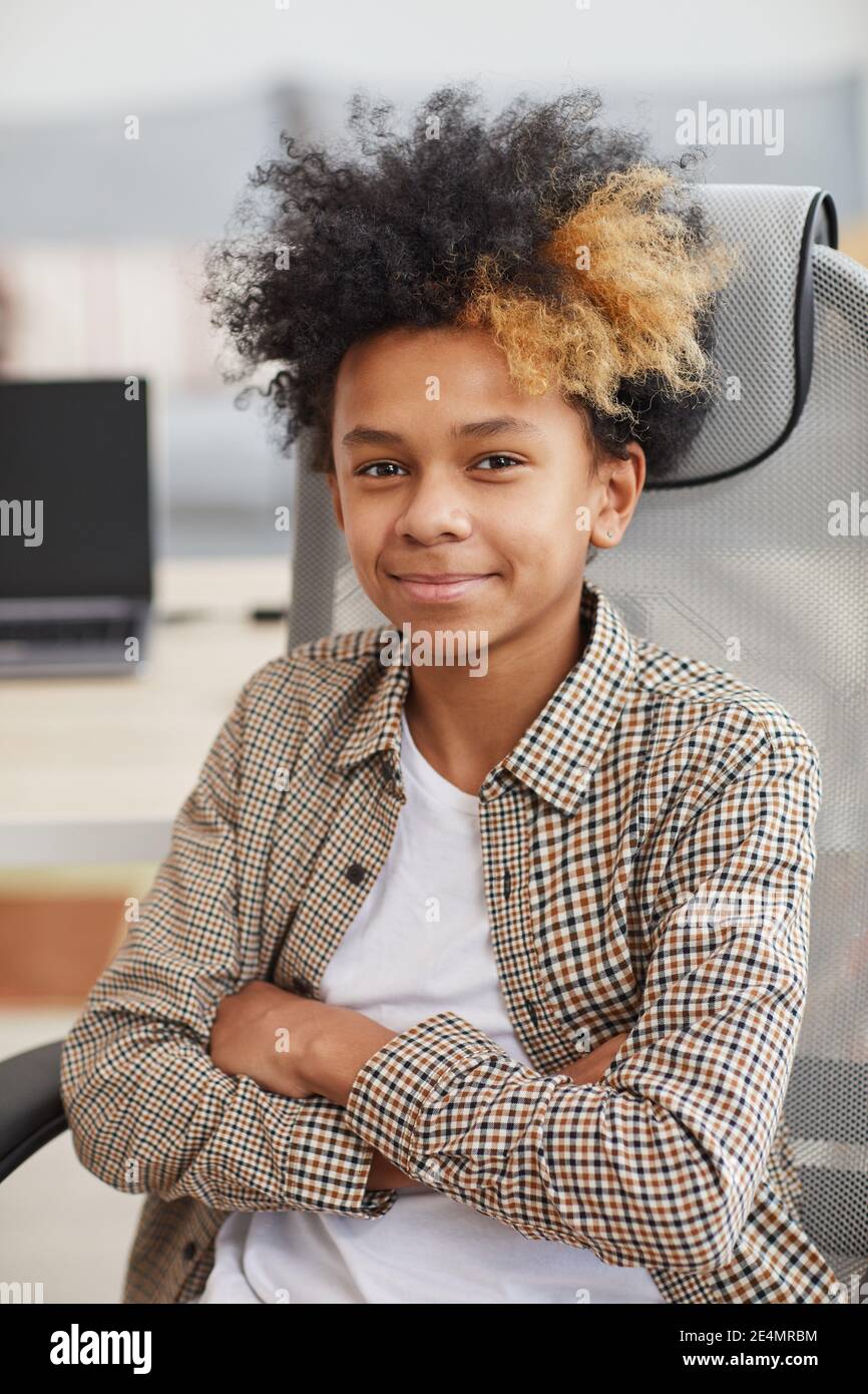 Ritratto verticale di ragazzo afro-americano adolescente sorridente alla macchina fotografica mentre seduto in sedia contro il computer in background Foto Stock