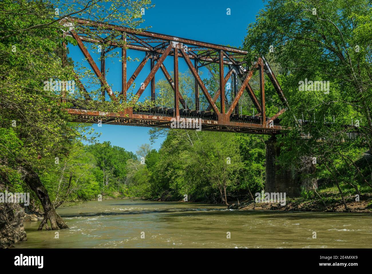 Un fiume in rapido movimento che scorre sotto il vecchio ferro arrugginito treno trenino con boschi circostanti su un cielo blu vibrante giorno in primavera Foto Stock