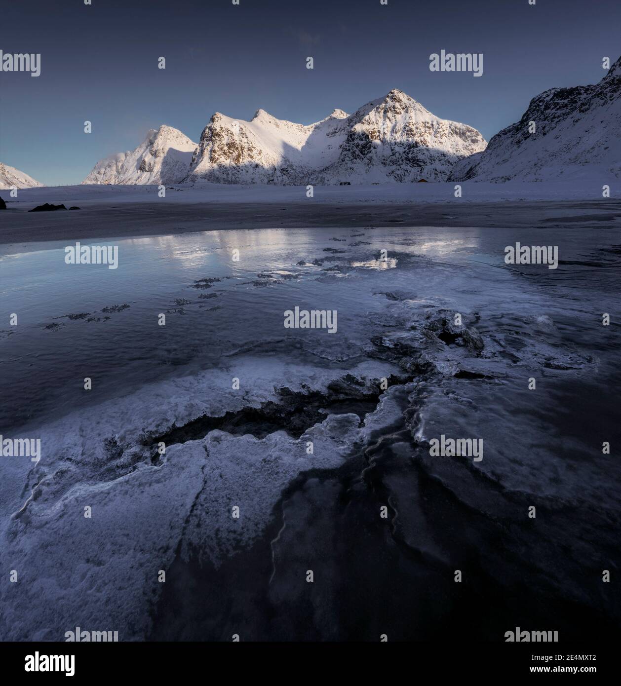 Tramonto invernale sulla spiaggia di Skagsanden, con interessanti texture nel ghiaccio speculare che riflettono le luminose montagne sullo sfondo. Foto Stock