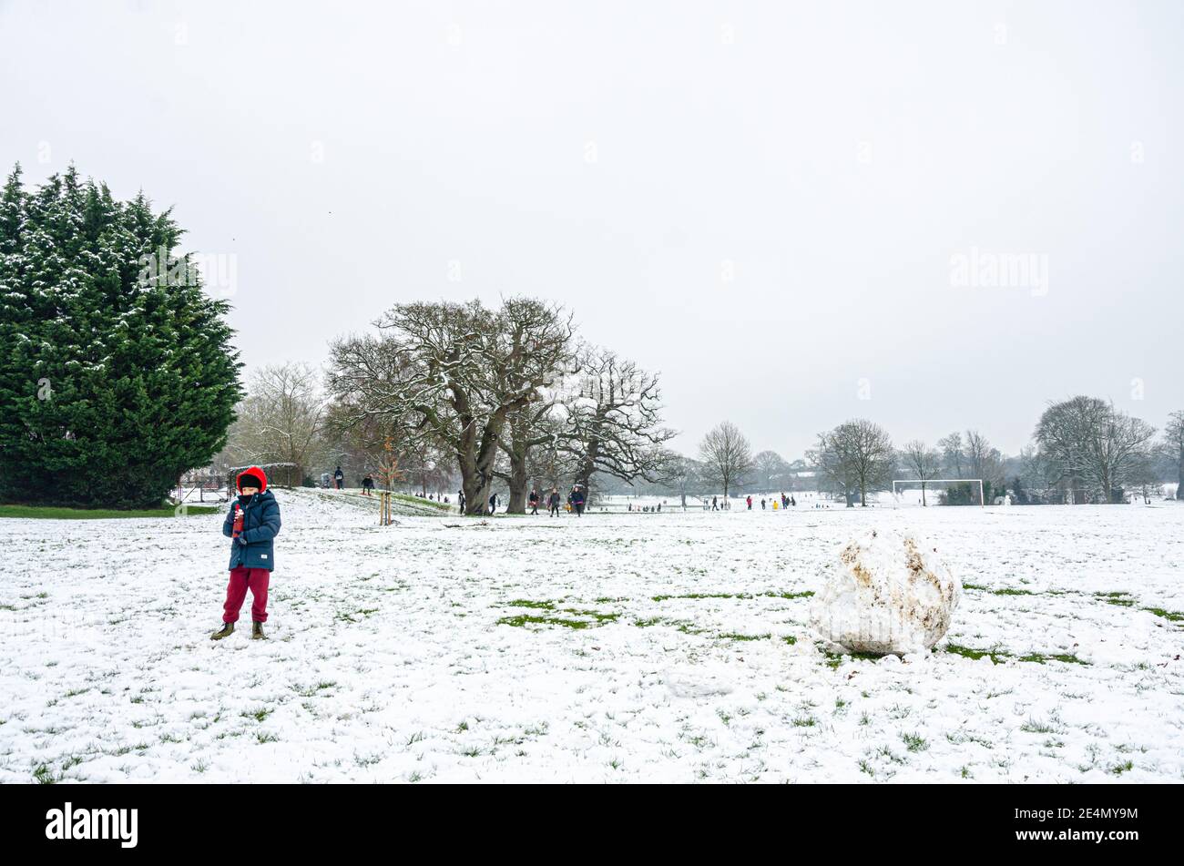 Un ragazzo che indossa un cappotto e un cappuccio rosso per una passeggiata trova un grande masso fatto di neve in Prospect Park a Reading, Berkshire, Regno Unito. Foto Stock