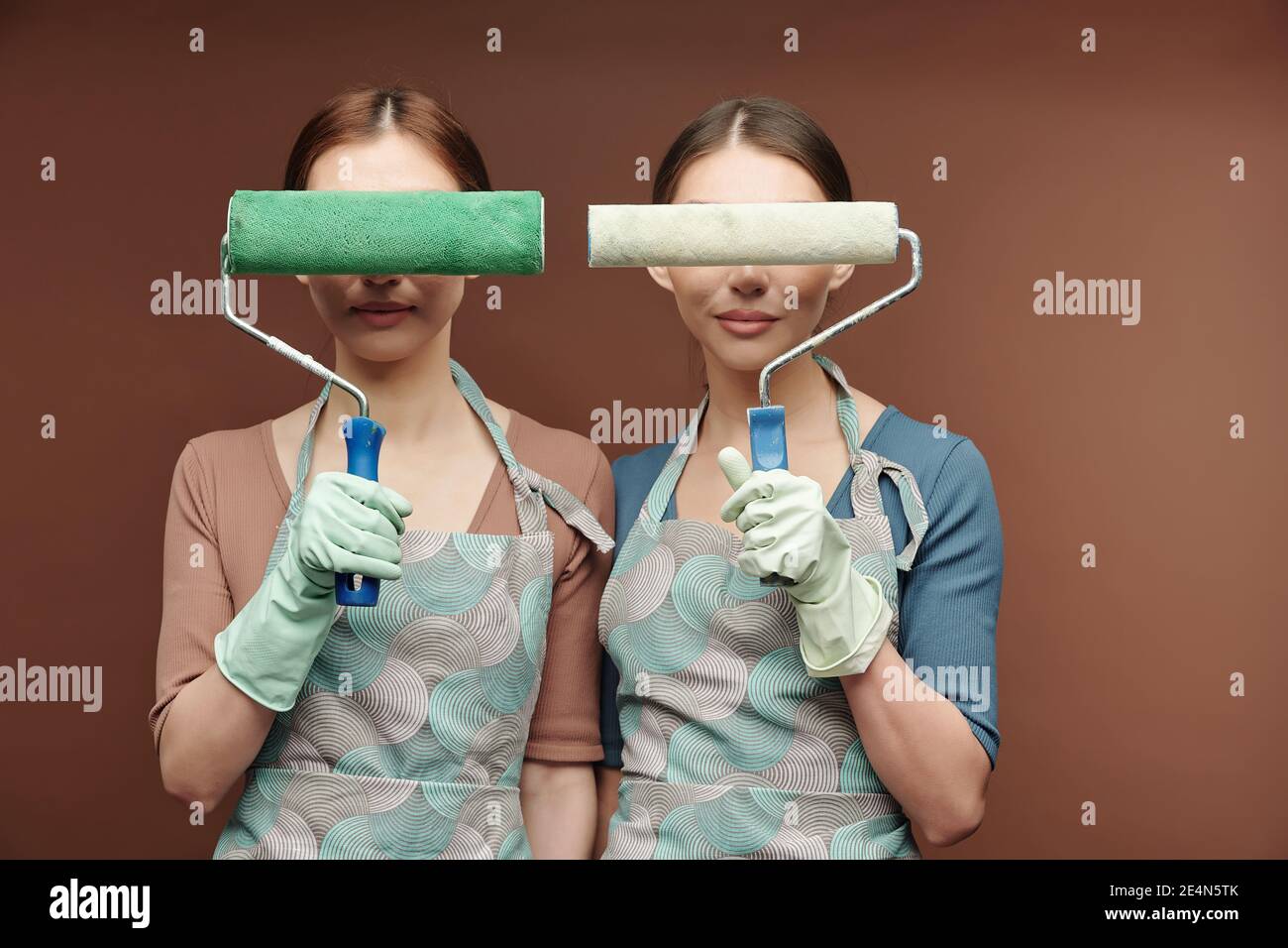 Gravi gemelle femminili giovani in grembiuli e guanti che tengono la vernice rulli per gli occhi mentre si è in piedi vicini l'uno all'altro davanti alla fotocamera Foto Stock