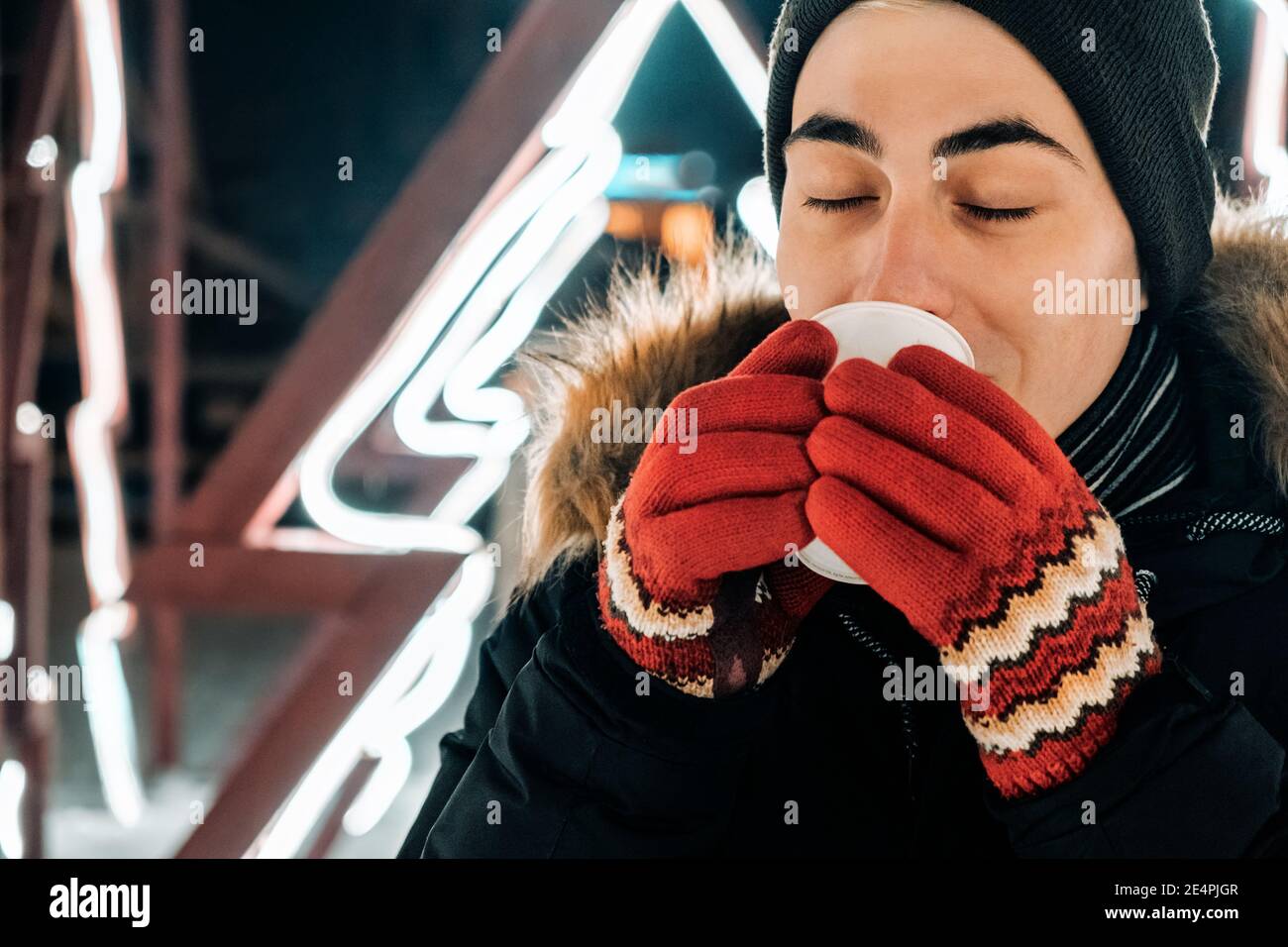 giovane uomo caucasico che beve cioccolata calda, caffè o tè in abiti invernali e guanti rossi su uno sfondo di retroilluminazione fluorescente Foto Stock