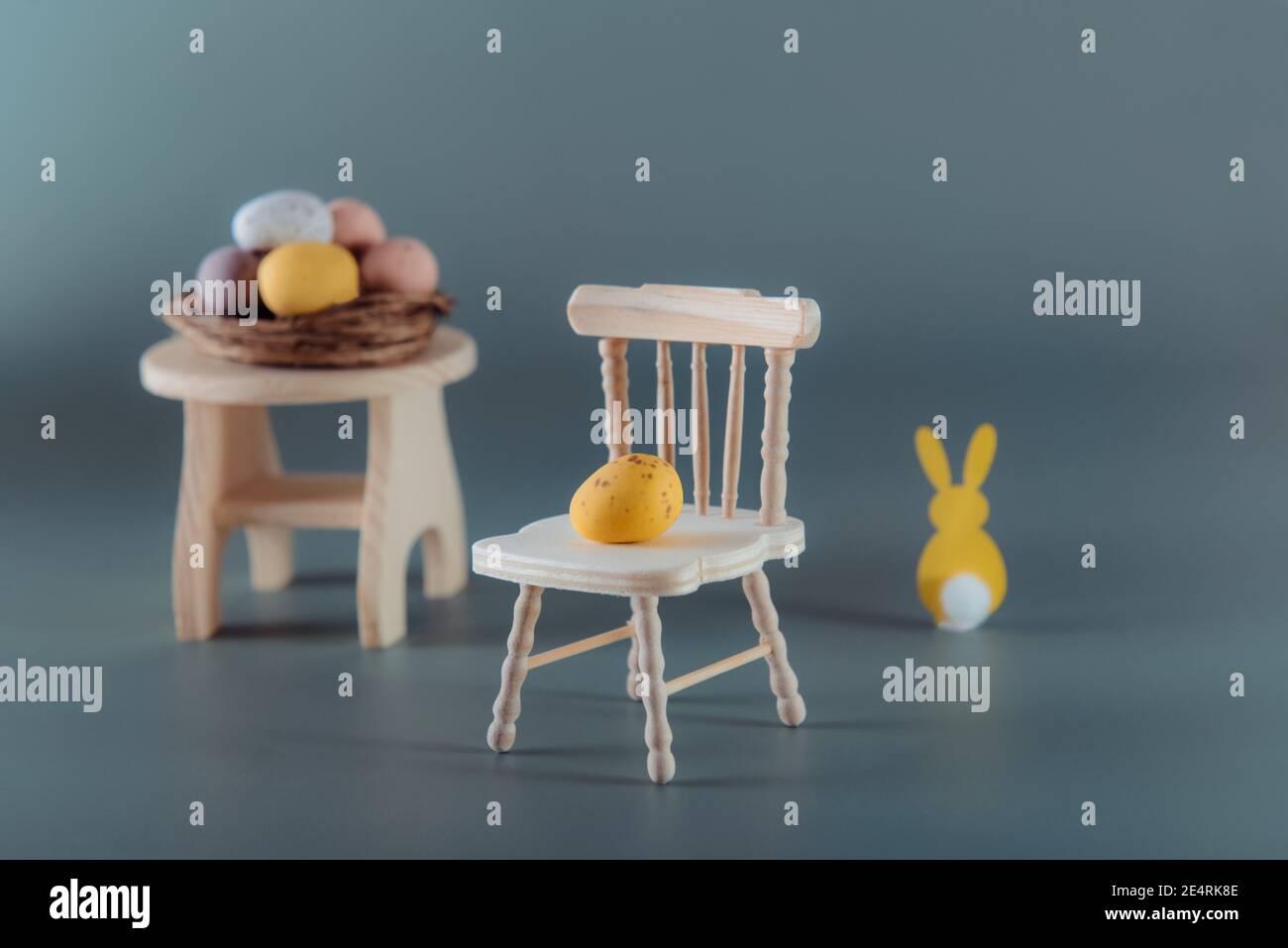 Composizione creativa di Pasqua di uovo di cioccolato giallo su una sedia in legno miniatura, coniglio coniglietto di pasqua e nido con uova caramelle sul tavolo sul muto Foto Stock