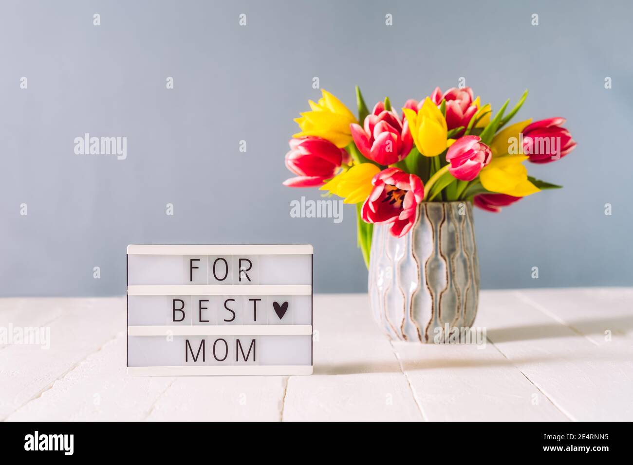 Scatola luminosa con per il miglior messaggio di mamma e bouquet di fiori di tulipano rosa e giallo in vaso su tavolo di legno bianco e sfondo grigio chiaro. Saluto festivo Foto Stock