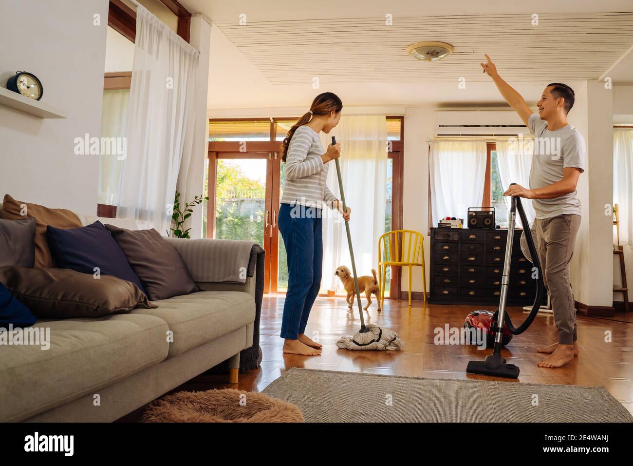 Felice giovane coppia di donne che danzano e cantano durante la pulizia con aspirapolvere e uomo che pulisci il pavimento soggiorno in casa moderna Foto Stock