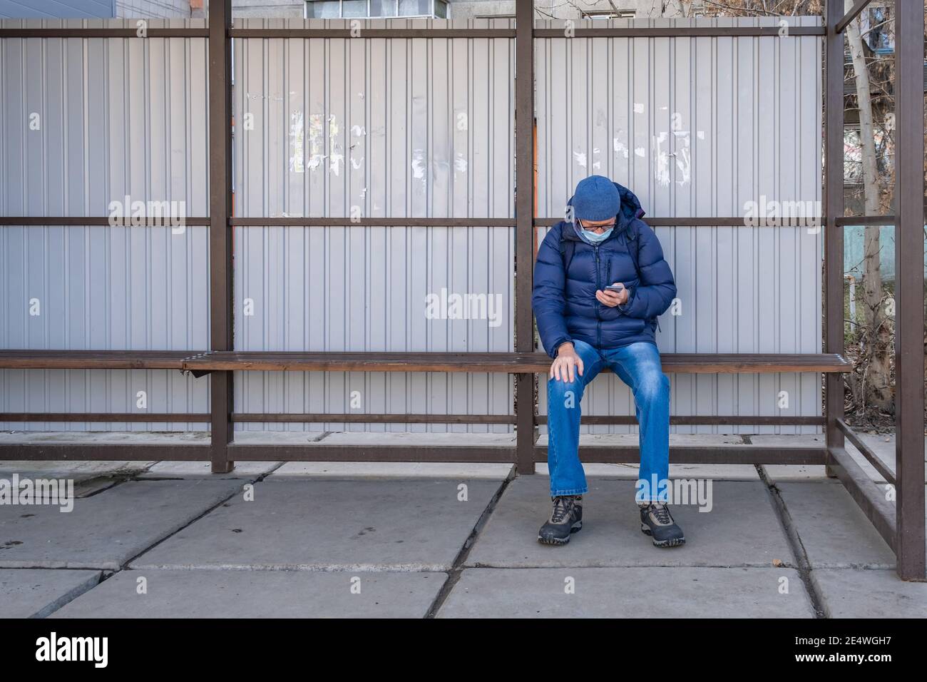 Un uomo maturo in maschera medica si siede in autunno ad una fermata di trasporto pubblico e attende un autobus, tram, filobus. Utilizzando lo smartphone, cerca informazioni Foto Stock