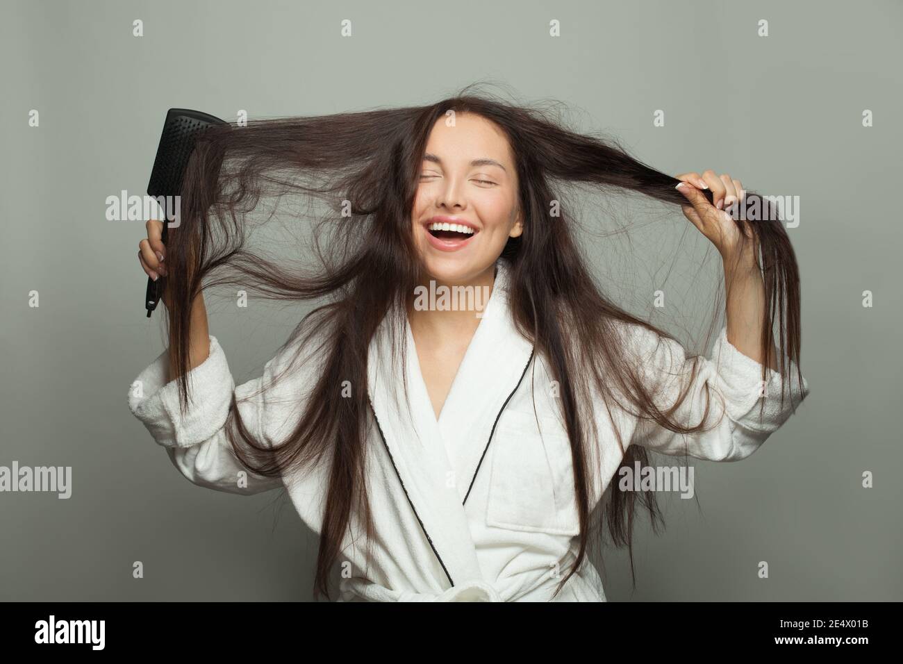 Felice giovane donna che combatte i suoi capelli aggrovigliati e sorridendo su sfondo bianco. Concetto di routine mattutina Foto Stock