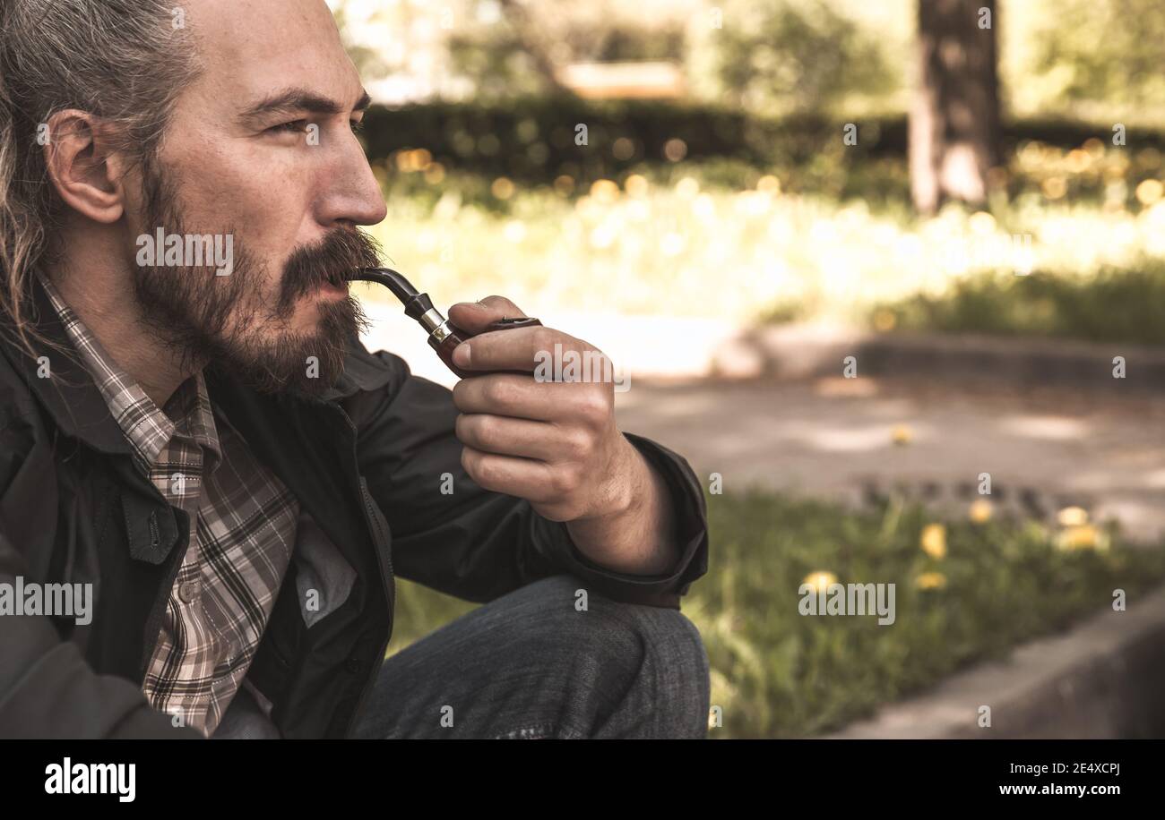 Brutale uomo bearded fuma una pipa nel parco estivo di giorno, primo piano ritratto Foto Stock